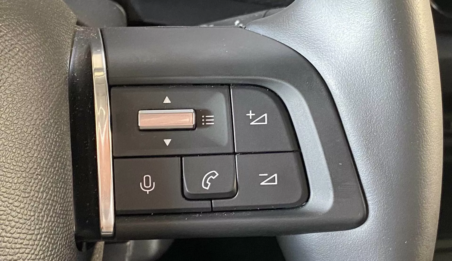 Vista aproximada de um painel de controle multifuncional no lado esquerdo do volante de um Citroën C4 2020 (e-C4) na perspectiva do motorista. O painel possui botões com símbolos para controle de volume, avanço de faixas, gerenciamento de chamadas telefônicas e um joystick para navegação no menu. O painel preto e os botões são projetados para feedback tátil e combinam com a estética do volante, que apresenta uma superfície texturizada e uma costura indicativa de material de alta qualidade.