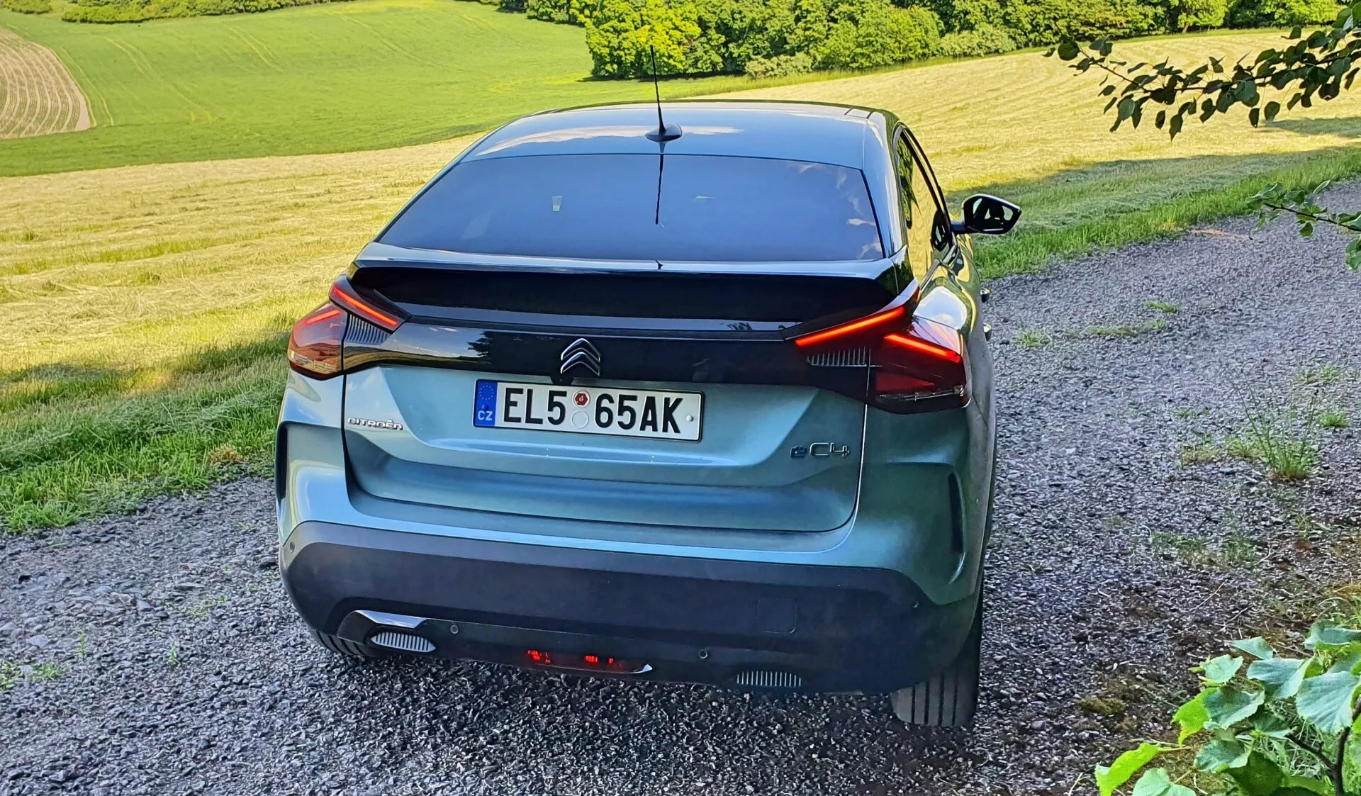 Vista traseira de um carro hatchback moderno capturado de um ângulo três quartos, enfatizando suas linhas elegantes, luzes traseiras elevadas e envolventes, placa de matrícula central, spoiler aerodinâmico e limpador traseiro. O veículo, com um acabamento metálico cinza ou azul, está equipado com sensores no para-choques. O carro está estacionado em um caminho de cascalho em um ambiente rural com grama e folhagem, fotografado no final da tarde ou início da noite, a partir da altura de uma pessoa de pé.