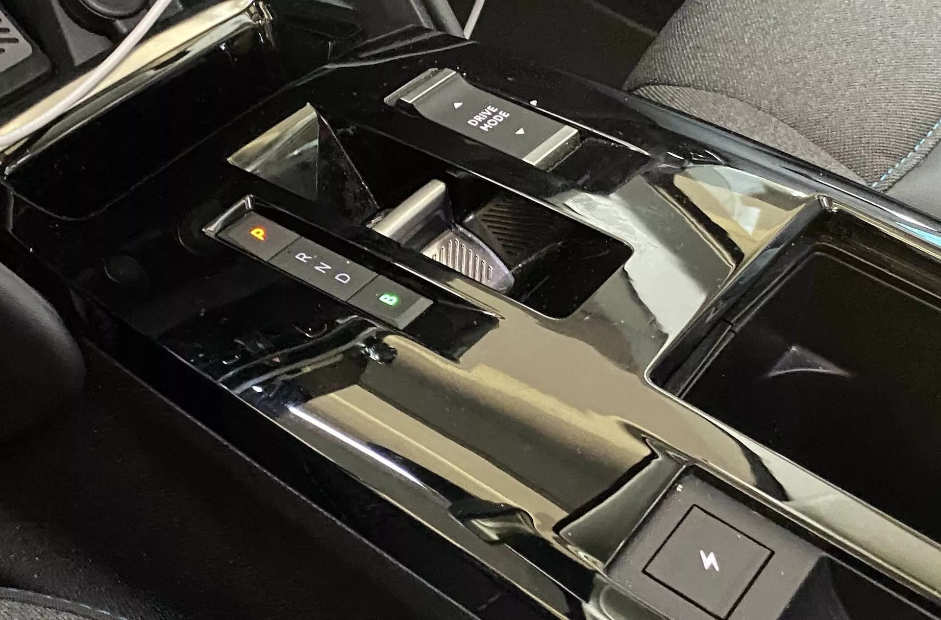 Vista aproximada da área do console central dentro de um Citroën C4 2020 (e-C4), destacando o seletor de marchas eletrônico, botões de controle de modos de condução, porta-copos, tampa do compartimento de armazenamento brilhante, interruptor do freio eletrônico de estacionamento, acabamento em material preto polido e interface de tela sensível ao toque parcialmente visível, sugerindo um interior de veículo de luxo e alta tecnologia.