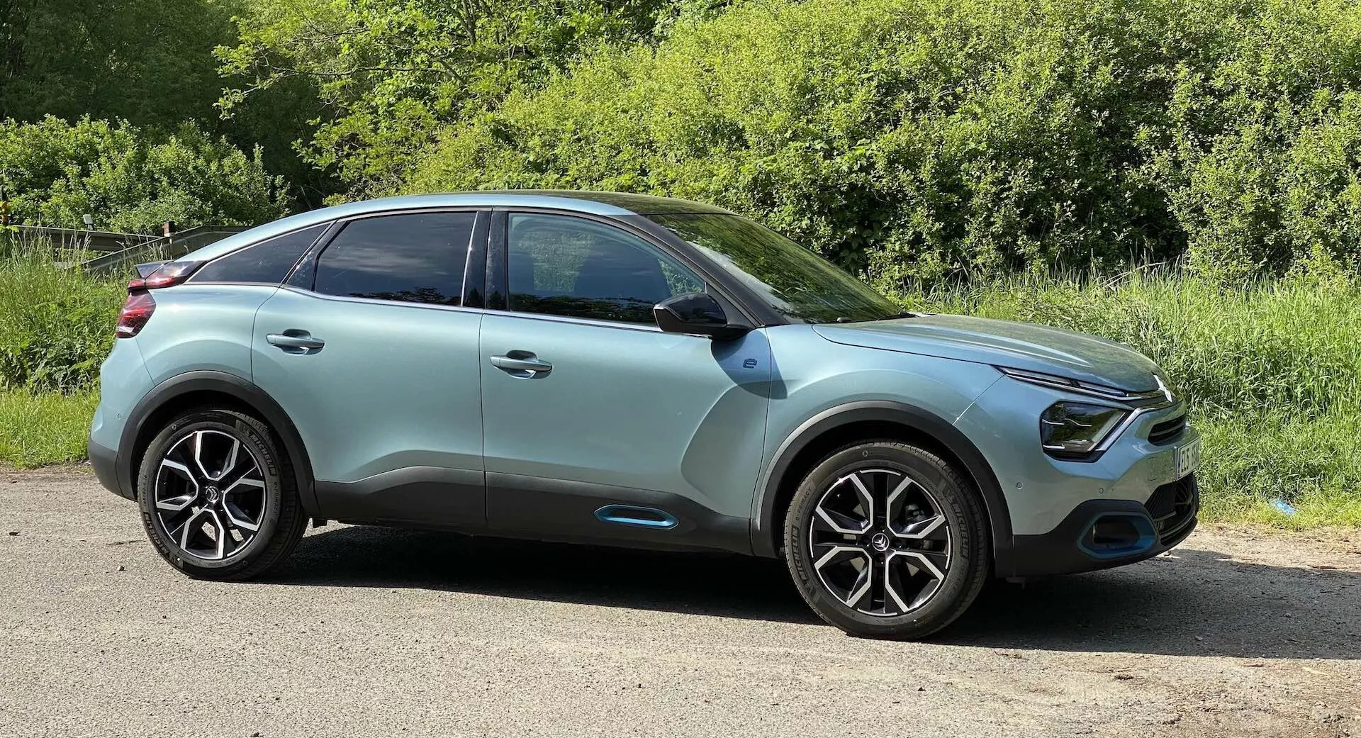 Perfil lateral do Citroen C4 2020 (e-C4), um SUV crossover moderno, tirado de um ângulo mais baixo. O carro está estacionado em uma superfície plana à beira da estrada com vegetação ao fundo, sugerindo um ambiente rural ou de parque. O elegante capô leva a uma grade dianteira curva ladeada por luzes diurnas de LED e possíveis faróis de neblina. A carroceria de dois tons em verde menta claro com adornos pretos mostra recursos avançados, como indicadores de direção integrados nos retrovisores laterais e sensores de entrada sem chave nas maçanetas das portas da mesma cor da carroceria. As grandes rodas de liga leve multiraio e os pneus de alto desempenho enfatizam a estética e o desempenho. A linha do teto suavemente inclinada, as janelas com vidro de privacidade e os possíveis sistemas avançados de assistência ao motorista indicam um foco na segurança, conforto e aerodinâmica. Capturada em luz natural com o carro posicionado a uma distância moderada, a imagem destaca o design exterior do veículo.