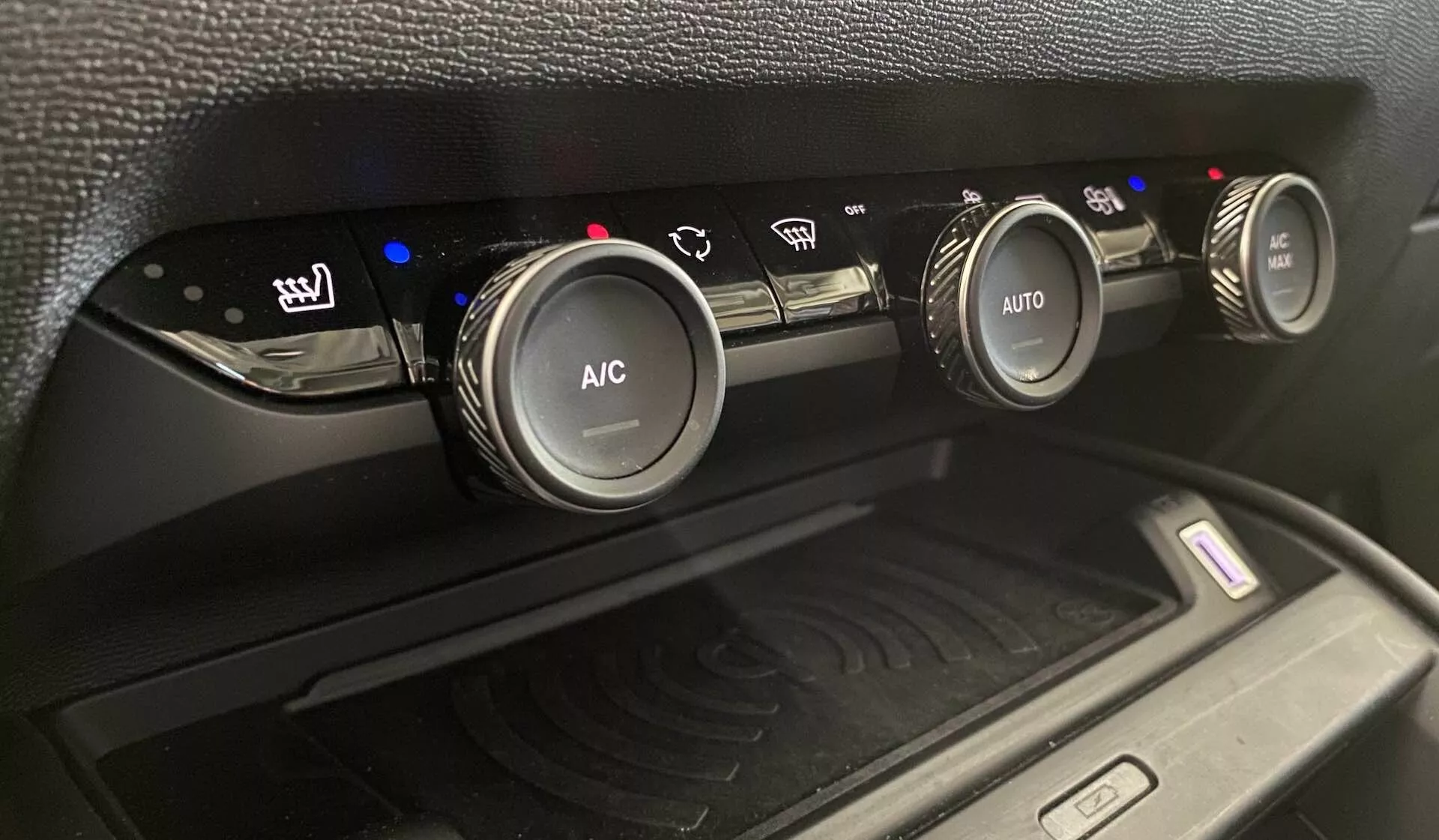 Close-up do sistema de controle climático do Citroen C4 2020 (e-C4), mostrando três botões de controle circulares no console central de um ângulo ligeiramente inclinado para cima. O botão à esquerda controla os assentos aquecidos com indicadores de nível de calor azuis, o botão central está rotulado como 'A/C' para o ar condicionado e o botão direito tem 'AUTO' para configurações automáticas de clima. Indicadores azuis e vermelhos refletem as configurações de temperatura. Abaixo dos botões, um compartimento apresenta uma almofada de carregamento sem fio com um símbolo de ondas de rádio, cercado por um acabamento preto fosco de plástico e uma textura padrão.