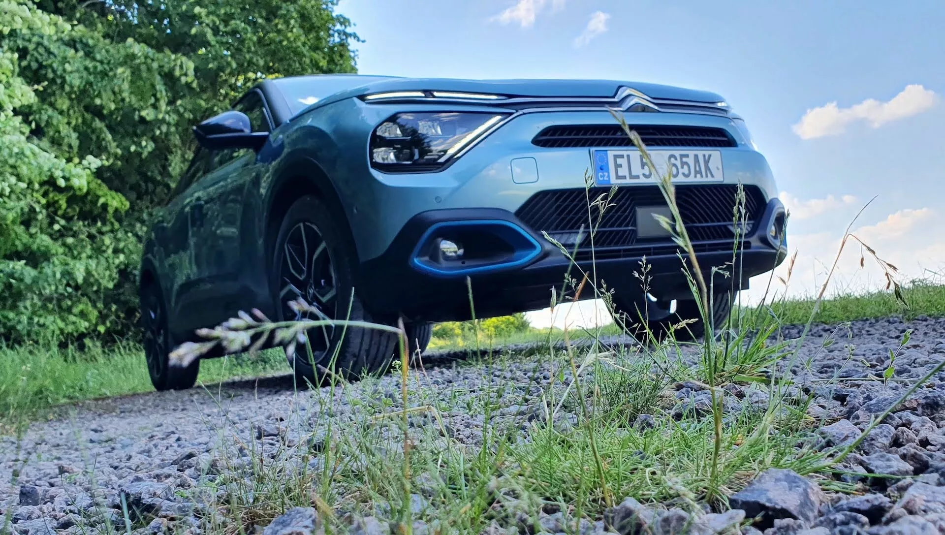 Vista frontal em ângulo baixo de um Citroën C4 2020 (e-C4) estacionado em um caminho de cascalho cercado por vegetação, mostrando o capô, a grade, o para-choque dianteiro, os faróis e uma parte das rodas dianteiras do carro. A imagem enfatiza a postura robusta do veículo e as características de design exterior, como a forma dos faróis, o padrão da grade, as entradas de ar e os contornos do capô. A luz do sol refletida na pintura destaca o apelo estético do carro. Potenciais tecnologias de iluminação avançadas e sensores de estacionamento também fazem parte das características tecnológicas visíveis.