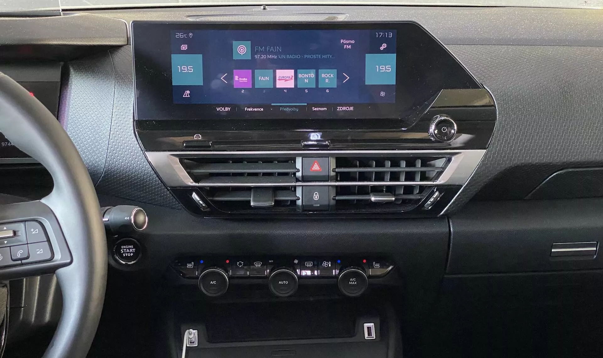 Vista interior do Citroën e-C4 2020 focalizada no painel a partir de uma posição central. A imagem mostra uma tela moderna de infoentretenimento com opções de navegação, mídia, conectividade telefônica e configurações do carro. Abaixo estão as saídas de ar horizontais com botões para o ar condicionado e outras funções, seguidas pelos controles de clima de zona dupla com botões de temperatura e um visor LCD. Parte do volante com controles montados e um botão de start-stop são visíveis, destacando a tecnologia moderna e o design ergonômico do carro.