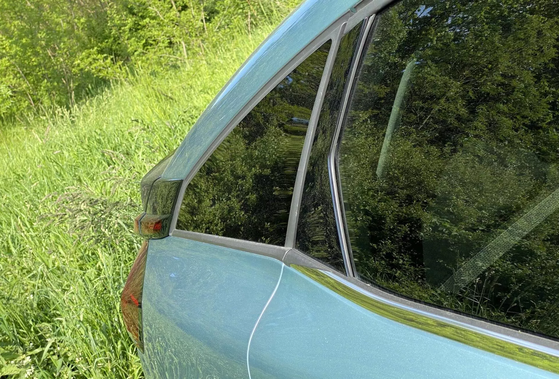 Vista aproximada do painel traseiro e janela do Citroën C4 2020 (e-C4). A foto, tirada de um ângulo oblíquo no lado traseiro do veículo, destaca a transição da janela traseira do passageiro para o para-brisa traseiro. A pintura azul claro metálica, a luz lateral laranja no pilar C, o contorno cromado e os reflexos nas superfícies de vidro são visíveis, destacando o design e o estado do carro.