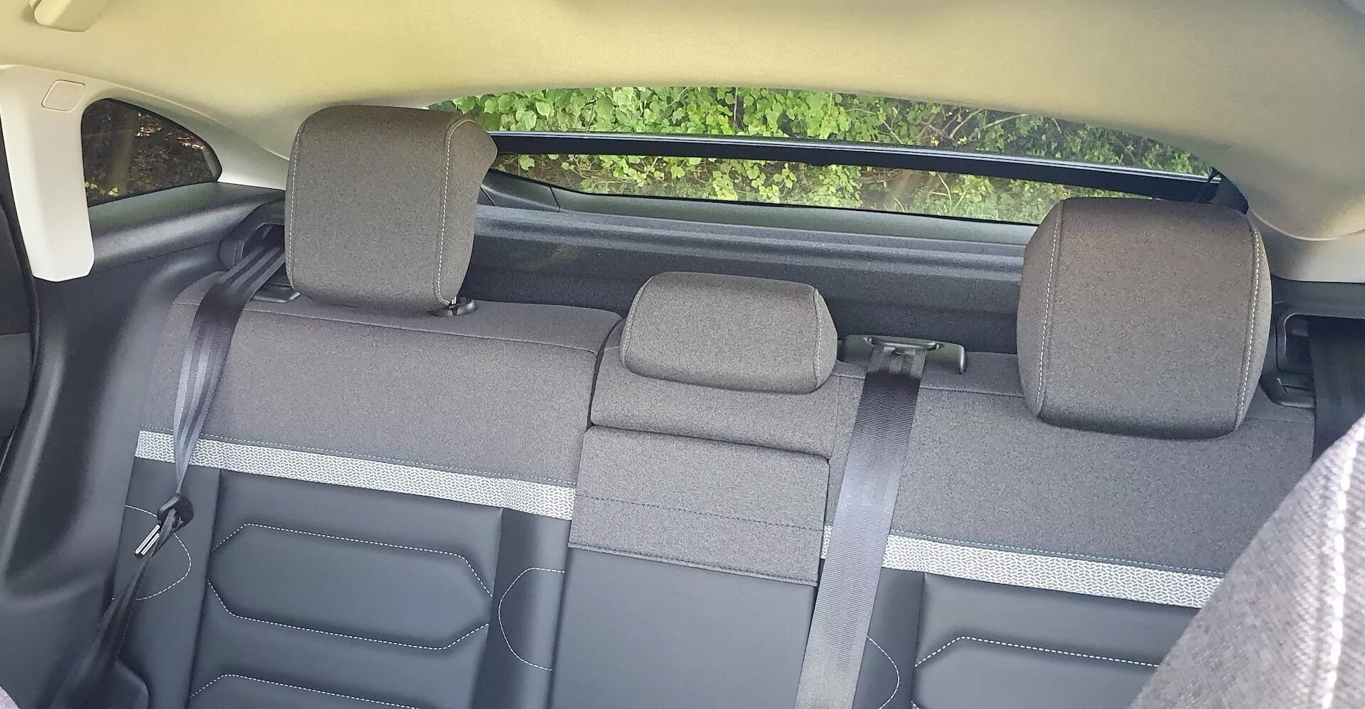Interior do Citroën C4 2020, focado nos assentos traseiros vistos dos assentos dianteiros. O banco traseiro, dividido em três seções com função de rebatimento 60/40, acomoda até três passageiros. O tecido da tapeçaria sugere um nível de acabamento padrão, com apoios de cabeça ajustáveis para os assentos exteriores. O esquema de cores é bicolor, com elementos plásticos visíveis nas laterais e nos alojamentos dos cintos de segurança. Nenhuma tecnologia avançada é visível, mas há características de segurança padrão, como os cintos de segurança.