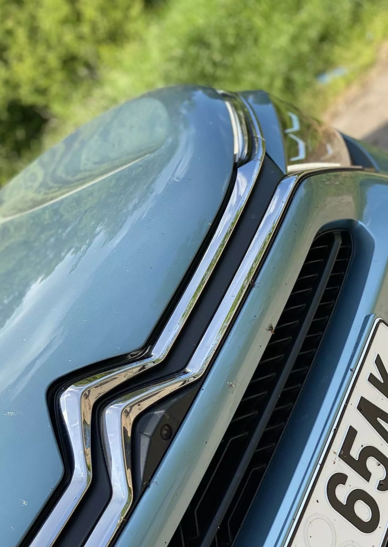 Close-up de um carro a partir de um ângulo frontal e ligeiramente superior, destacando a grade frontal com uma peça de cromo orientada verticalmente e lâminas horizontais, emblema destacado da marca no topo, e design esculpido do capô com contornos elevados. Sujeira ou pólen visível no capô indica um ambiente ao ar livre.