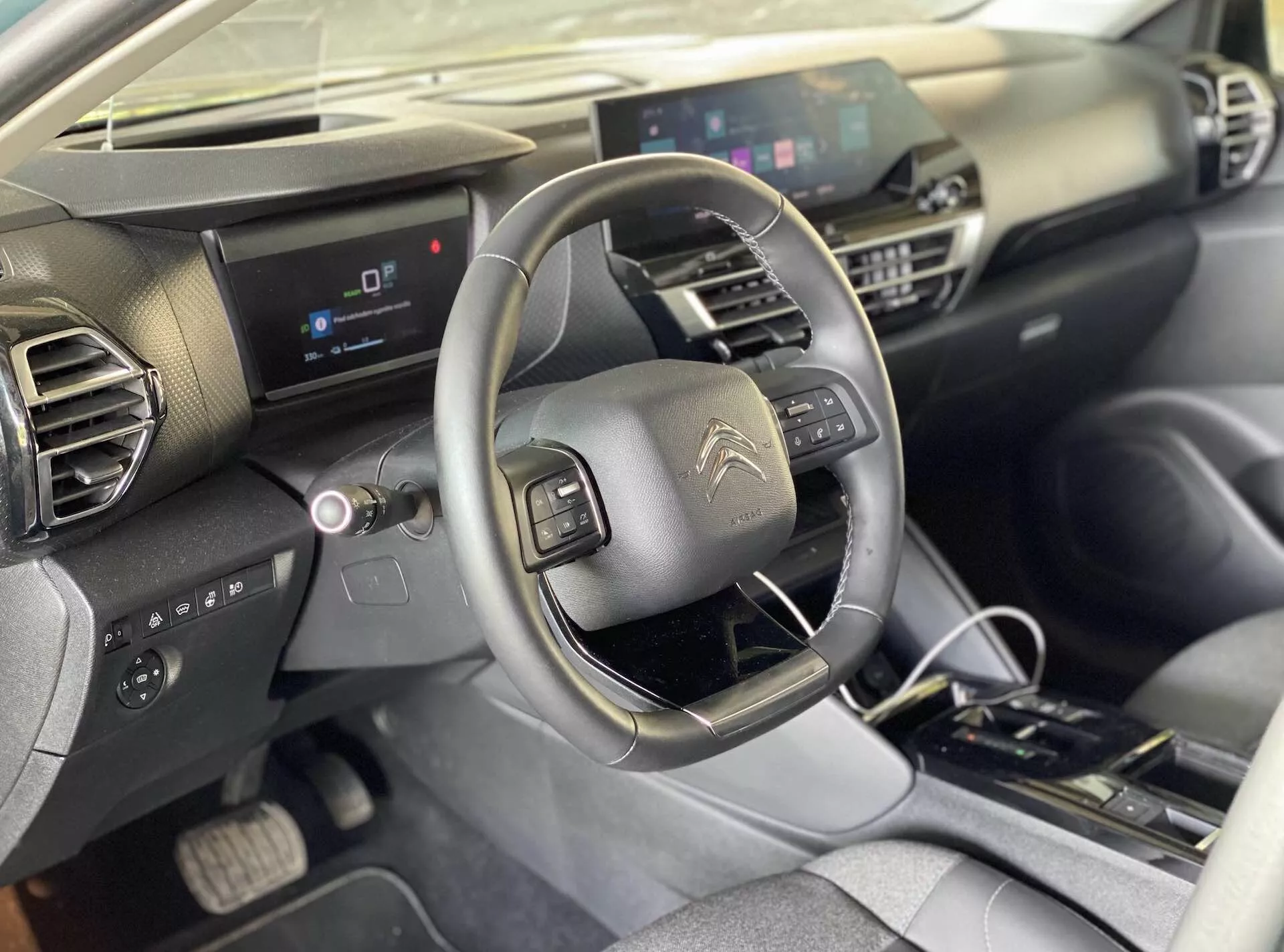 Vista interior de um Citroën e-C4 2020 a partir da perspectiva do passageiro do banco da frente, capturando o banco do motorista, console central, volante com controles integrados, painel de instrumentos digital e tela central de infotainment. O painel apresenta um design geométrico com materiais escuros e claros. O console central inclui controles físicos para o ar condicionado e um seletor de marchas automático.