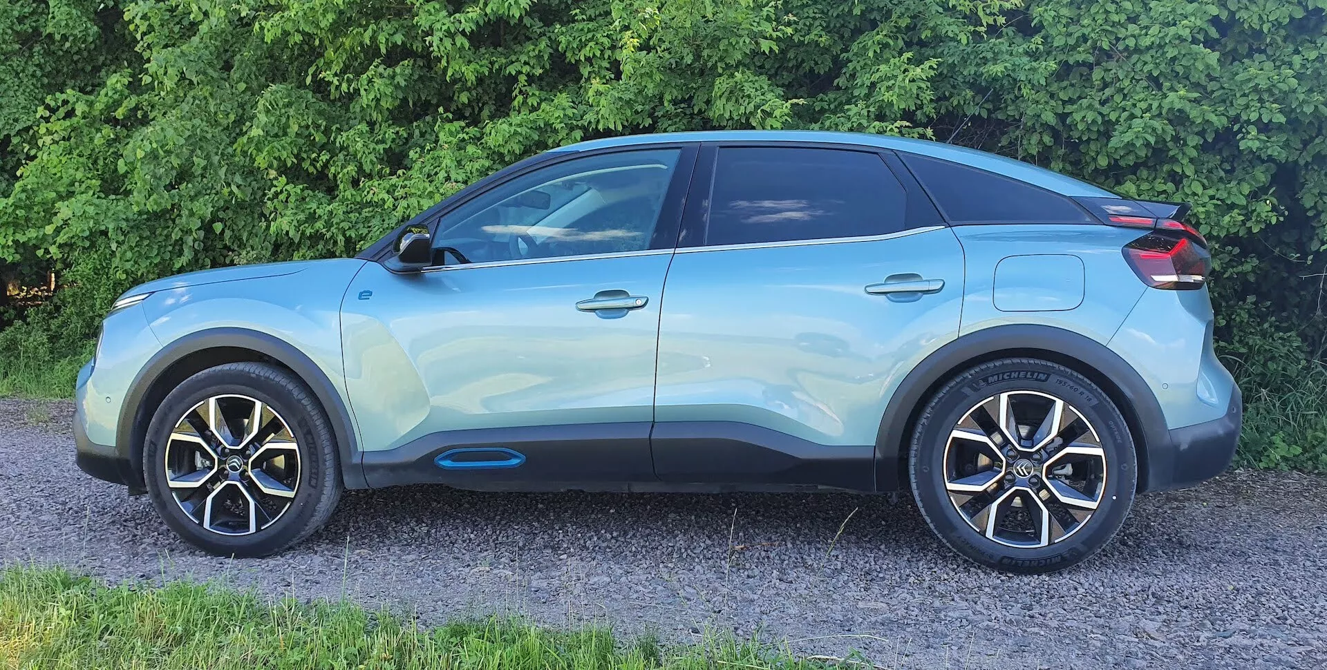 Vista de perfil lateral de um Citroen C4 (e-C4) crossover SUV moderno de 2020. O veículo azul claro está fotografado de um ângulo lateral-frontal, destacando detalhes como acabamento brilhante, rodas de liga, espelhos laterais da cor da carroçaria, linhas de caráter marcantes, acabamento preto das janelas, vidros traseiros escurecidos, maçanetas das portas embutidas com entrada sem chave e uma linha de teto inclinada com um possível spoiler de teto e trilhos de teto. A imagem mostra claramente a roda dianteira e o para-lama, mas exclui a grade frontal, o para-choque e os faróis.