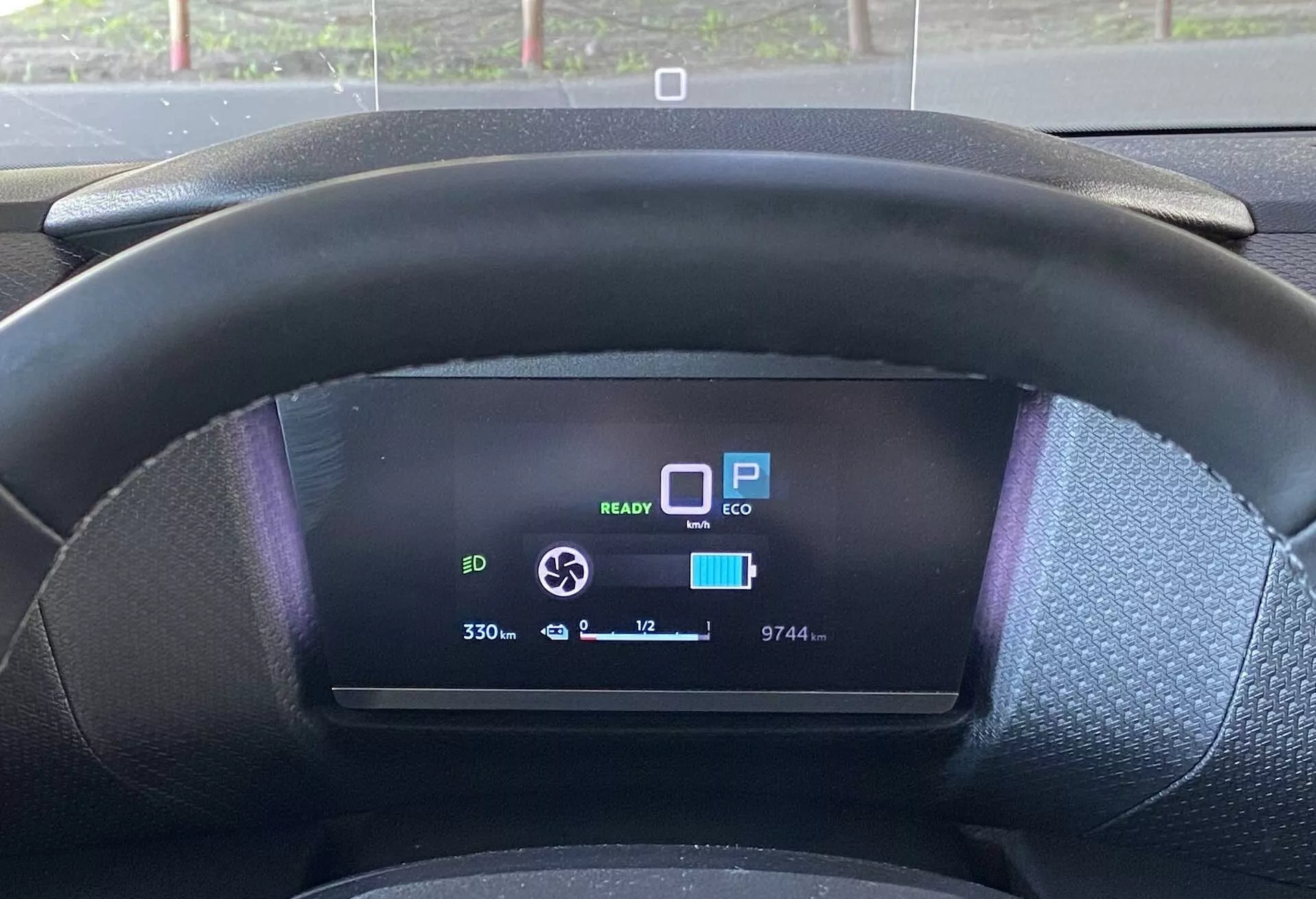 Vista aproximada do banco do condutor de um Citroën C4 2020 (e-C4) mostrando o volante texturizado preto e o painel de instrumentos digital. O volante cobre ligeiramente a parte inferior do display digital, que indica várias funcionalidades: ícone de prontidão EV 'READY,' 'P' para estacionar, modo 'ECO,' um ícone de bateria quase totalmente carregada, indicador de alcance mostrando '330,' odômetro lendo '9744,' temperatura externa '+12°C' e um ícone de seta circular possivelmente para ajuste da força de frenagem regenerativa.