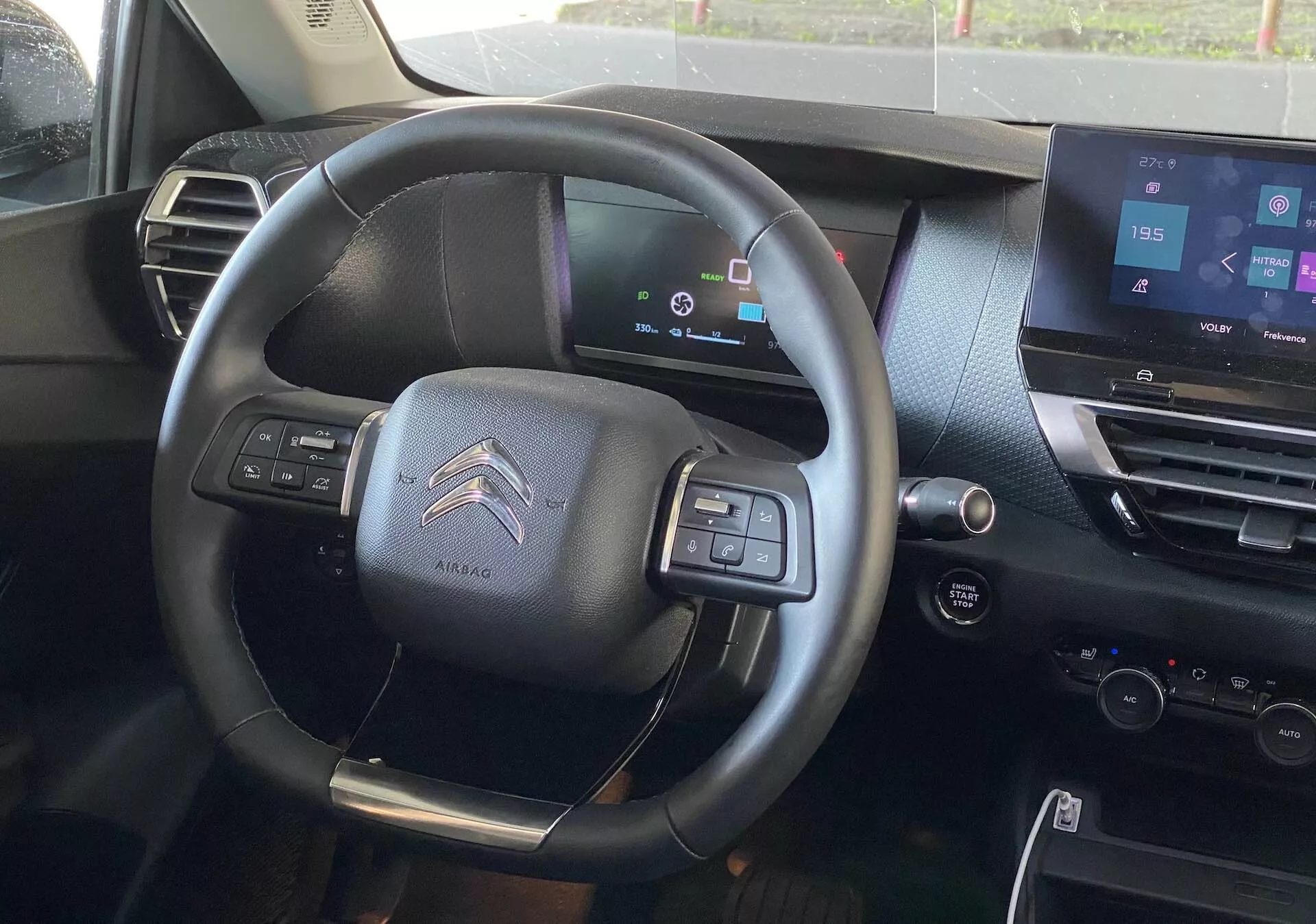 Vista do interior do Citroën C4 2020 (e-C4) a partir do assento do condutor, com um volante multifuncional de três raios centrado com anotação de airbag, painel de instrumentos digital mostrando status do veículo, sistema de infotainment com tela sensível ao toque na consola central, design de interior em tons escuros, botões de controle para áudio e conectividade de telefone, pás de mudança sugerindo uma transmissão semi-automática e controles na porta do condutor para vidros elétricos e ajustes dos espelhos laterais.