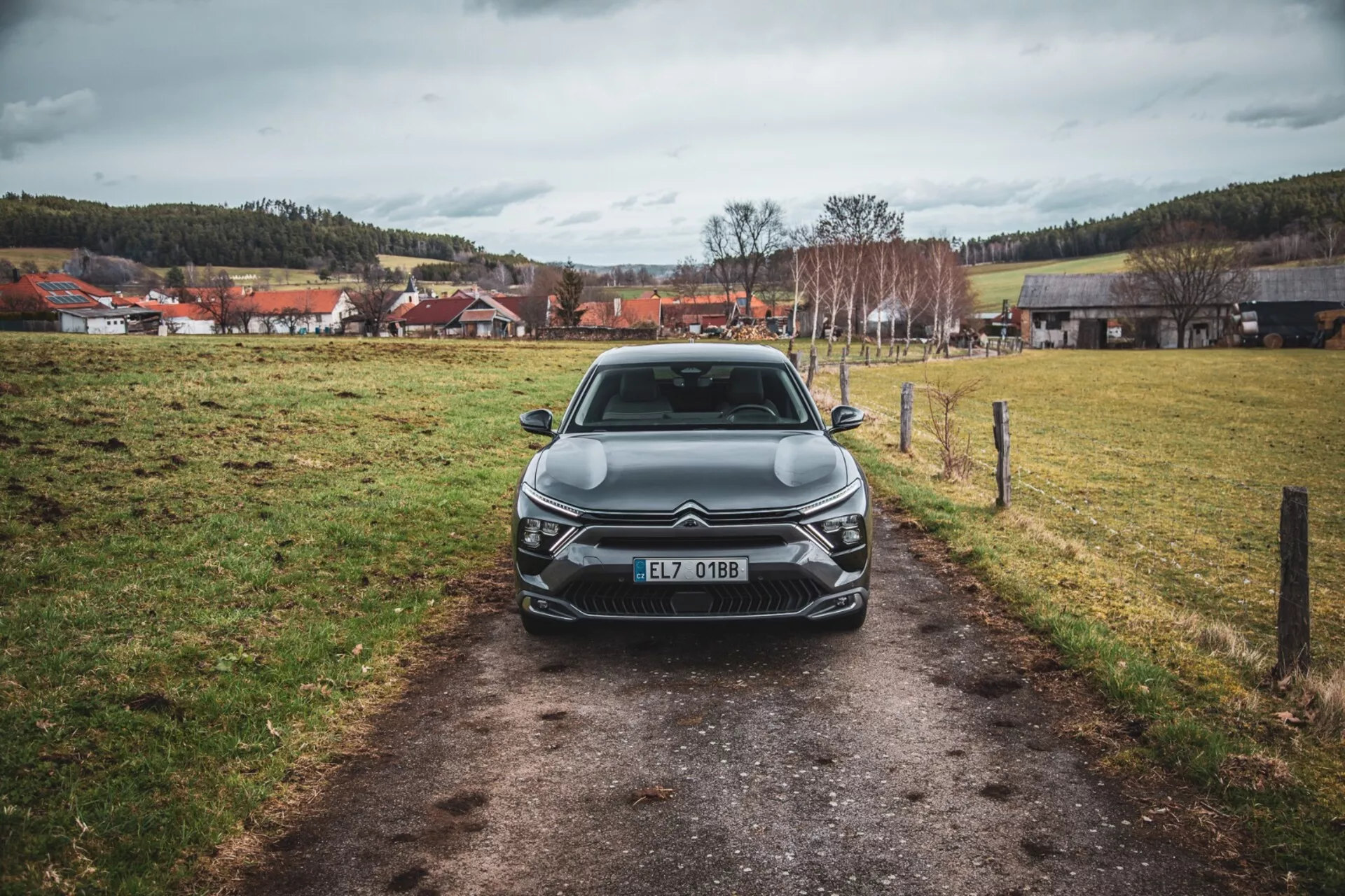 Vista frontal de um Citroën C5-X 2022 num caminho estreito em um campo gramado sob um céu nublado, mostrando seu design elegante, grelha intricada com emblema, unidades de faróis LED angulares, linhas esculpidas no capô e elementos adicionais de iluminação frontal, registrado na República Tcheca.