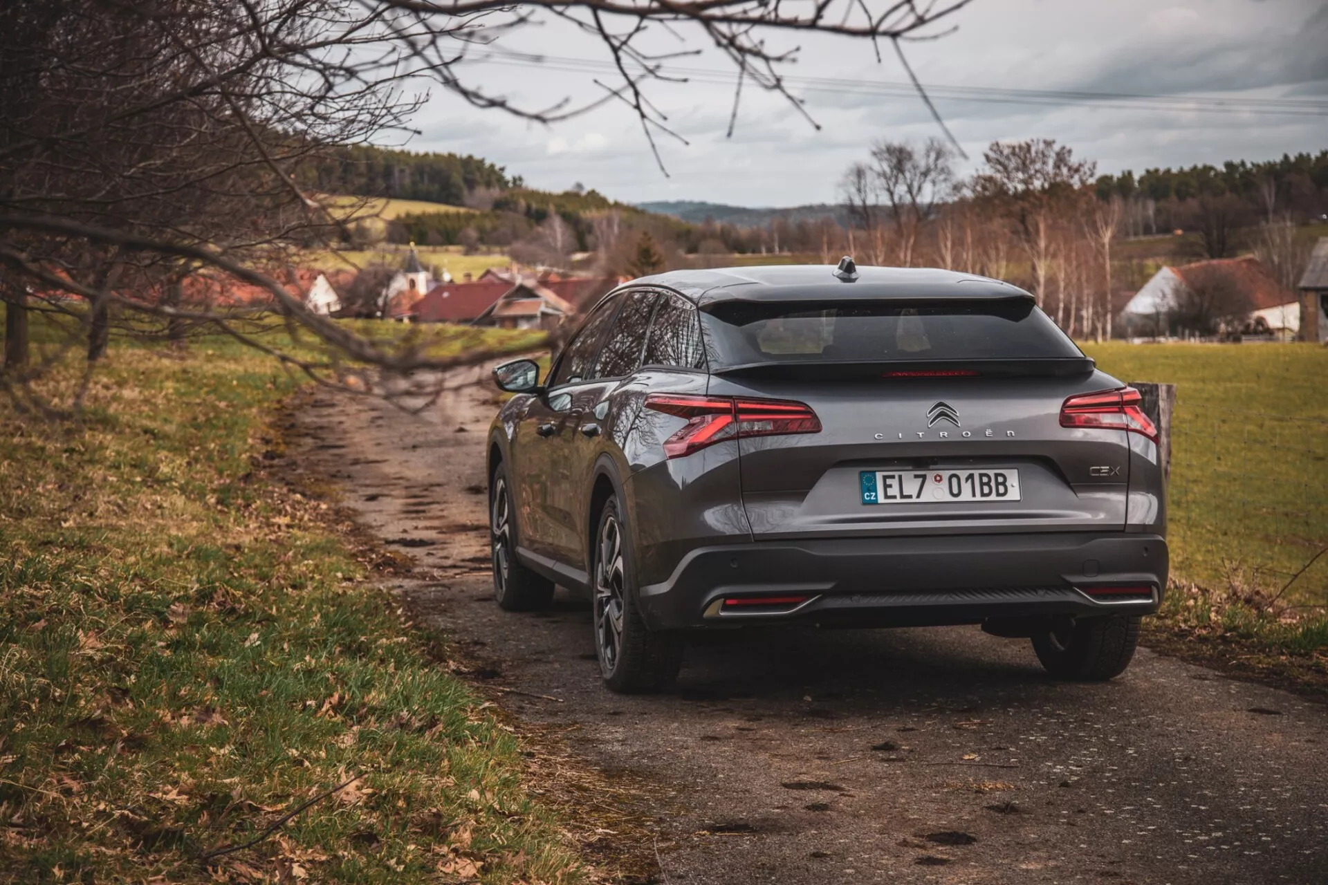 Vista traseira do hatchback Citroën C5-X 2022 em um caminho de terra com pano de fundo de uma pequena aldeia, mostrando luzes traseiras, para-choque traseiro, ponteiras de escape duplas, um emblema indicando que é um modelo Citroën, e um acabamento cinza metálico sob iluminação nublada