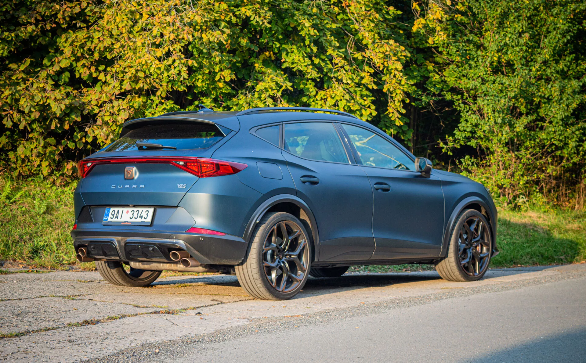 SUV crossover Cupra Formentor 2020 cinza escuro fosco preto estacionado no asfalto, exibindo vista traseira de três quartos que enfatiza design tipo cupê, aerodinâmica esportiva, lanternas traseiras LED, aerofólio traseiro, escapamentos duplos e grandes rodas de liga leve em um cenário suburbano