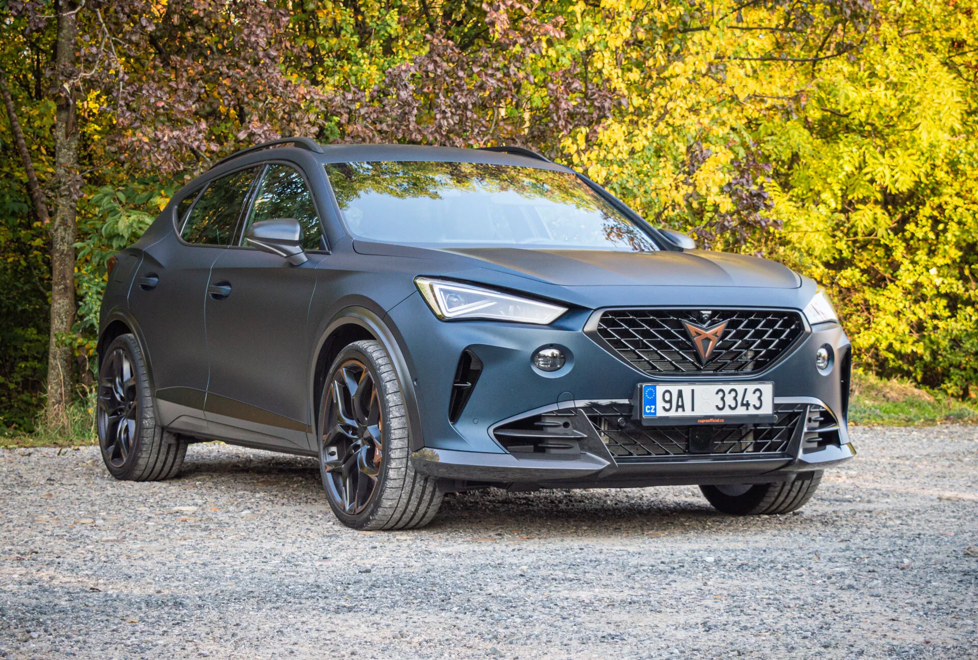 Cupra Formentor de 2020 estacionado numa superfície de cascalho com folhagem de outono ao fundo, exibindo seu exterior cinza fosco, grade distintiva, faróis de LED e acabamento esportivo preto