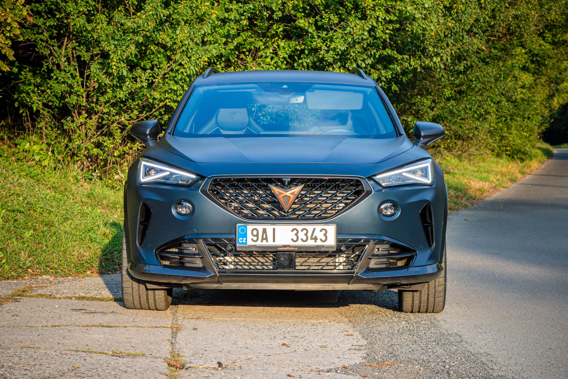 Vista frontal moderna do Cupra Formentor 2020 em uma estrada pavimentada com luz natural diurna e cenário rural, destacando grade agressiva, faróis LED nítidos, contornos dinâmicos do capô e características de tecnologia avançada