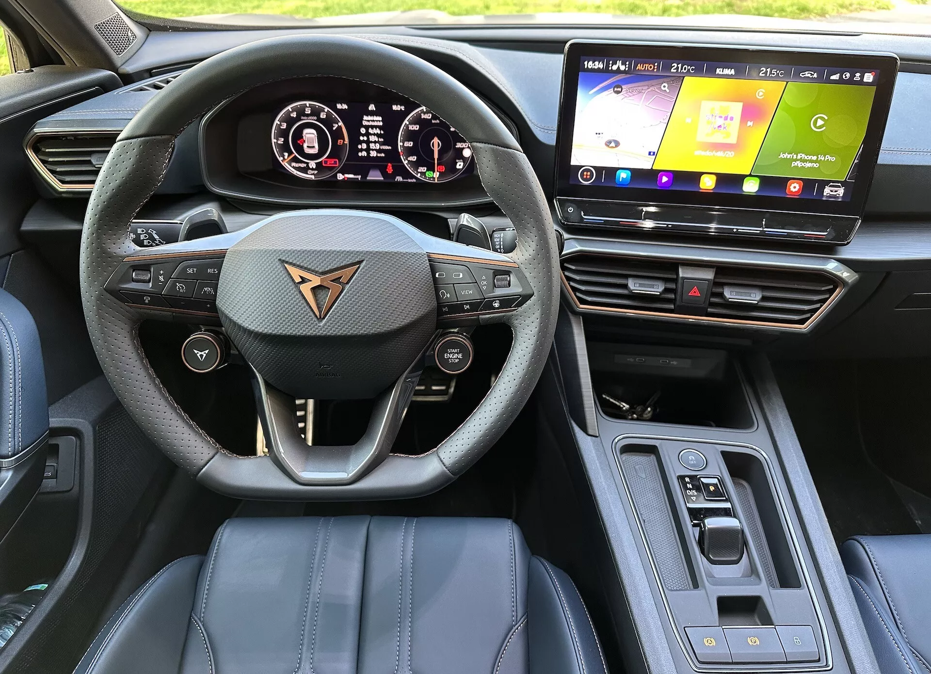 Vista interior do Cupra Formentor 2020 a partir da perspectiva do motorista mostrando volante multifuncional revestido a couro com o logótipo da marca do carro, cluster de instrumentos digital, sistema de infotainment touchscreen, materiais premium no painel com costura de destaque, seletor de marchas automático, e funcionalidades tecnológicas avançadas à luz natural