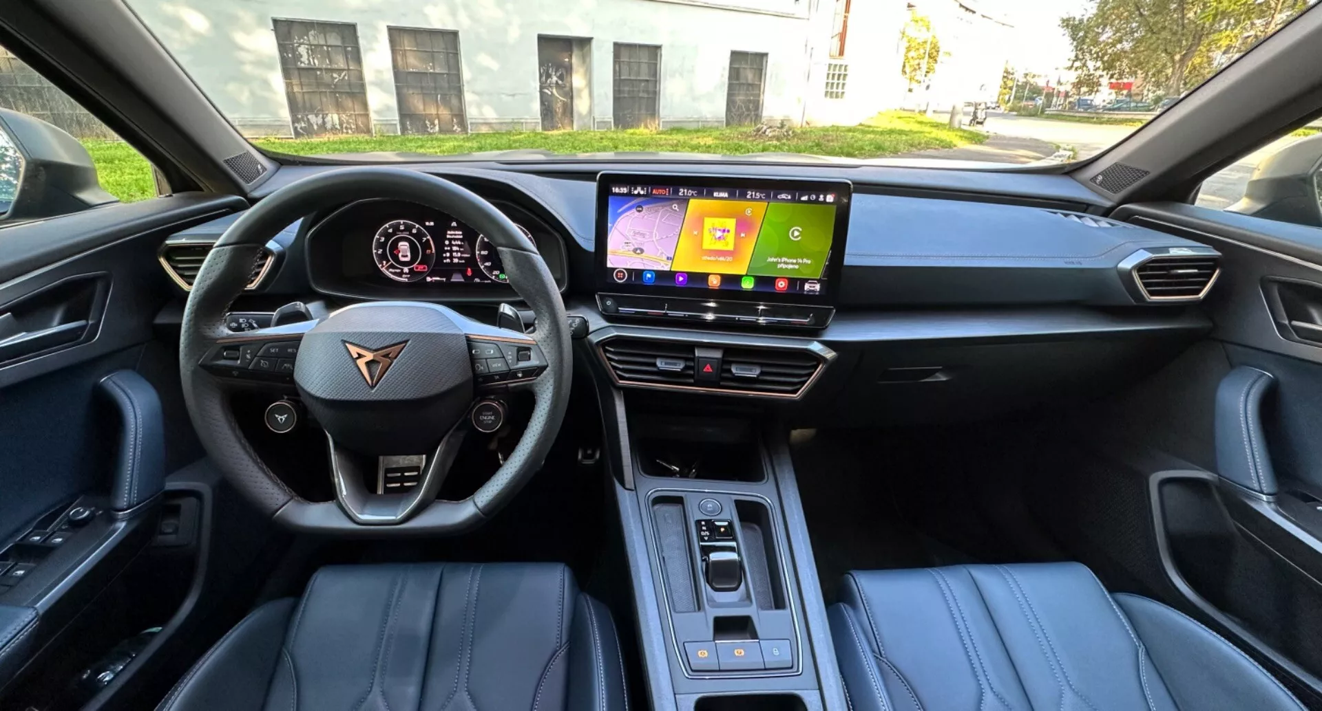 Vista interior de um CUPRA Formentor 2020 a partir do assento do motorista mostrando um volante de fundo plano com botões de multimídia e controle de cruzeiro, um painel digital exibindo velocidade e RPM, uma tela de infotainment central, selecionador de marchas automático elegante, assentos revestidos em couro com acento em duas cores, e iluminação da cabine durante o dia.
