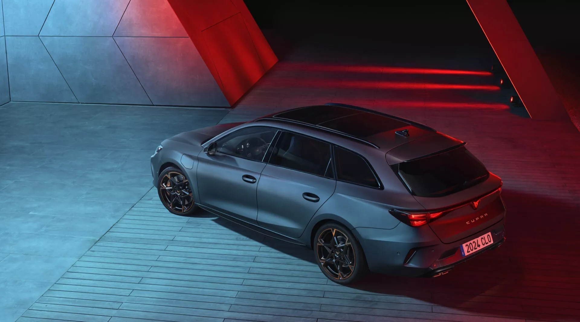 Vista em ângulo elevado de um Cupra Leon 2024 (1ª geração facelift) tirada à noite, mostrando a traseira e o lado direito do sportback. O carro está iluminado por luzes direcionais vermelhas dramáticas que criam uma atmosfera elegante e futurista. Apresenta um acabamento fosco, luzes traseiras LED iluminadas e rodas de liga aerodinâmicas. O veículo está estacionado em uma superfície industrial com padrões, sugerindo movimento. As características tecnológicas visíveis incluem um difusor traseiro e uma antena curta na parte traseira para tecnologias de comunicação.
