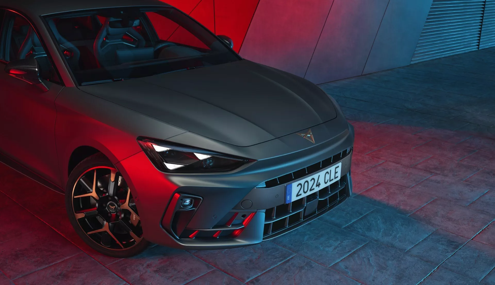 Vista de ângulo baixo frontal do Cupra Leon 2024 (primeira geração com facelift), destacando os recursos de design com uma grade proeminente, faróis elegantes com luzes diurnas de LED, estilização esportiva do para-choque com entradas de ar e rodas de liga leve orientadas para desempenho com pinças de freio de alto desempenho. O acabamento metálico da pintura e a iluminação ambiente vermelha dramática sugerem um veículo de alto desempenho, provavelmente fotografado no chão de uma sala de exposições.