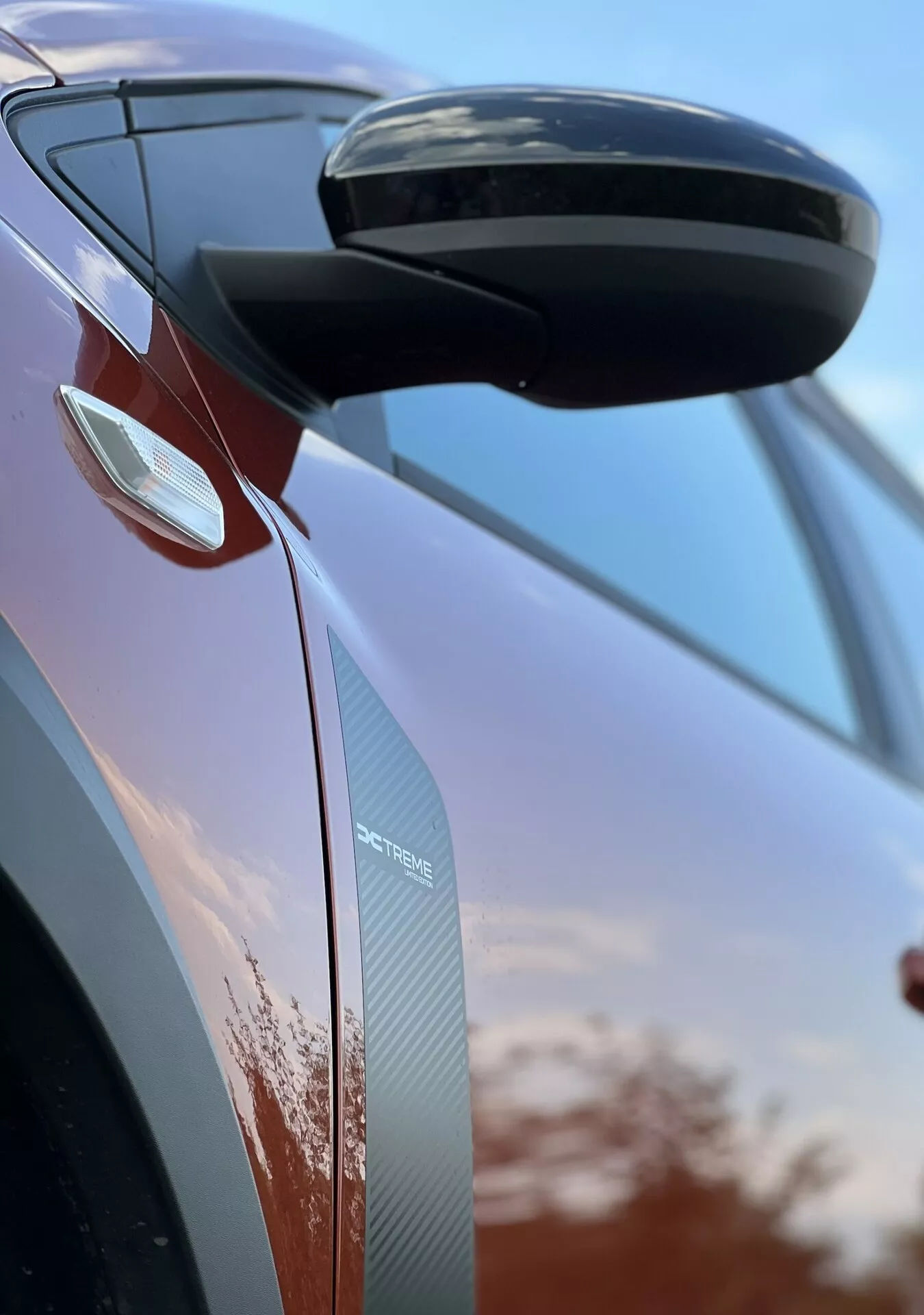 Detalhe em close-up do Dacia Jogger 2022 focando no espelho retrovisor lateral e no design da porta, exibindo características aerodinâmicas, sinais de seta integrados, e texturas de fibra de carbono contra um fundo de céu