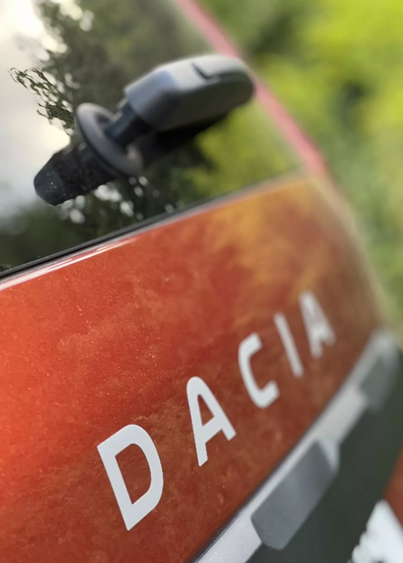 Vista aproximada do painel traseiro do Dacia Jogger 2022 com o nome DACIA em letras maiúsculas brancas contra uma pintura metalizada laranja-vermelha com manchas e sujeira, sugerindo uso e durabilidade ao ar livre, situado em um ambiente natural com um espelho lateral desfocado indicando características modernas do espelho.