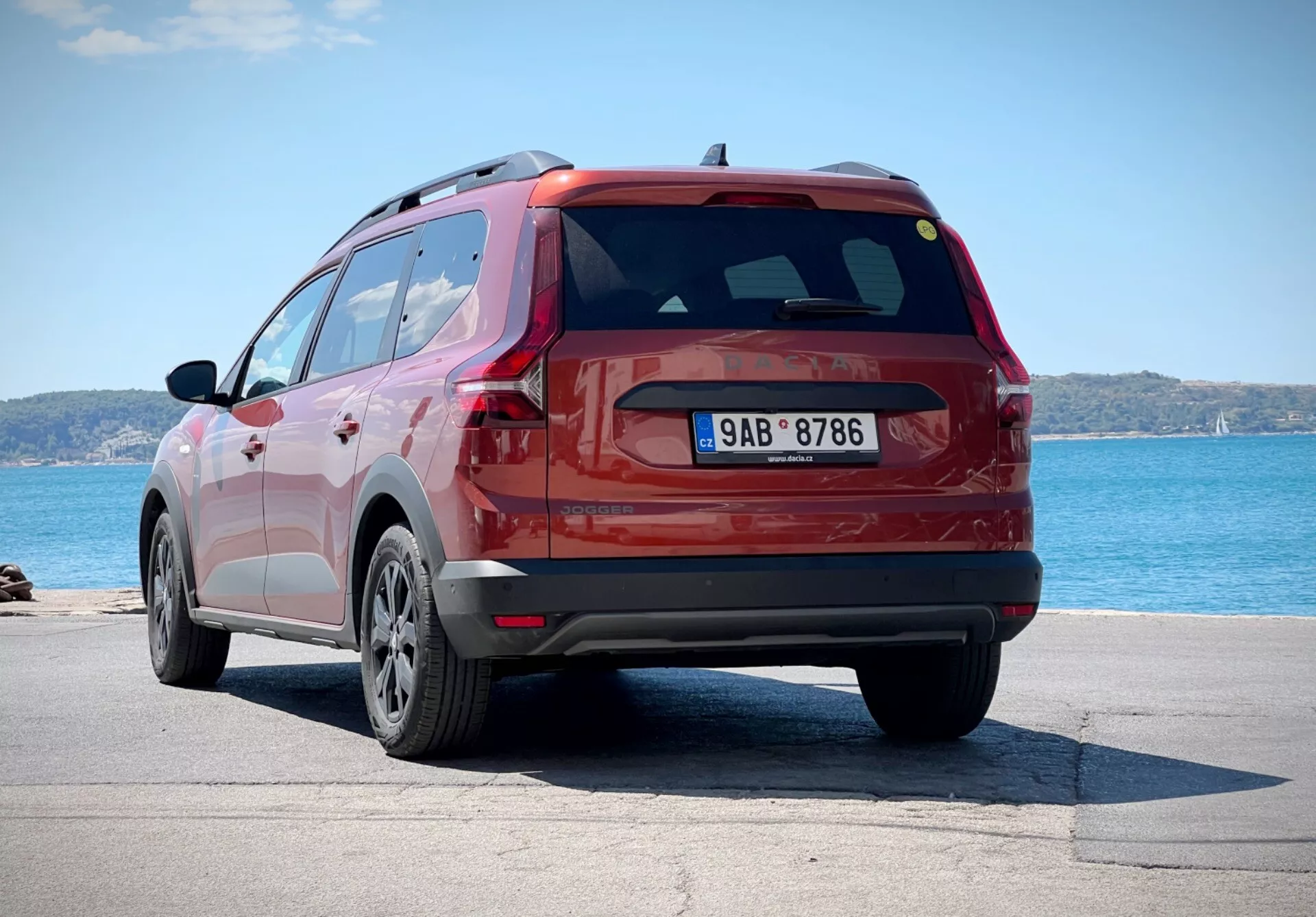 Dacia Jogger 2022 em vermelho, capturado de um ângulo traseiro de três quartos mostrando o lado e o design do portão traseiro do SUV, estacionado perto de um corpo de água com o horizonte ao fundo, destacando suas rodas de liga leve de múltiplos raios, luzes traseiras LED e spoiler traseiro aerodinâmico