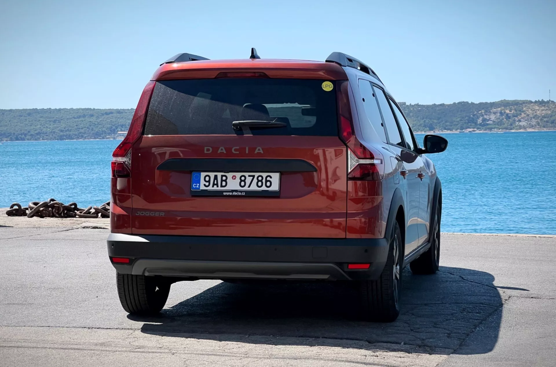 Dacia Jogger 2022 vermelho fotografado de um ângulo traseiro de três quartos, mostrando suas luzes traseiras, limpador de para-brisa traseiro, emblema, para-choque e placa contra um fundo de frente para a água com águas azuis e costa distante, destacando seu design hatchback/SUV com potenciais sensores de estacionamento e uma visão da porta traseira do passageiro, painel de quarto traseiro e roda.