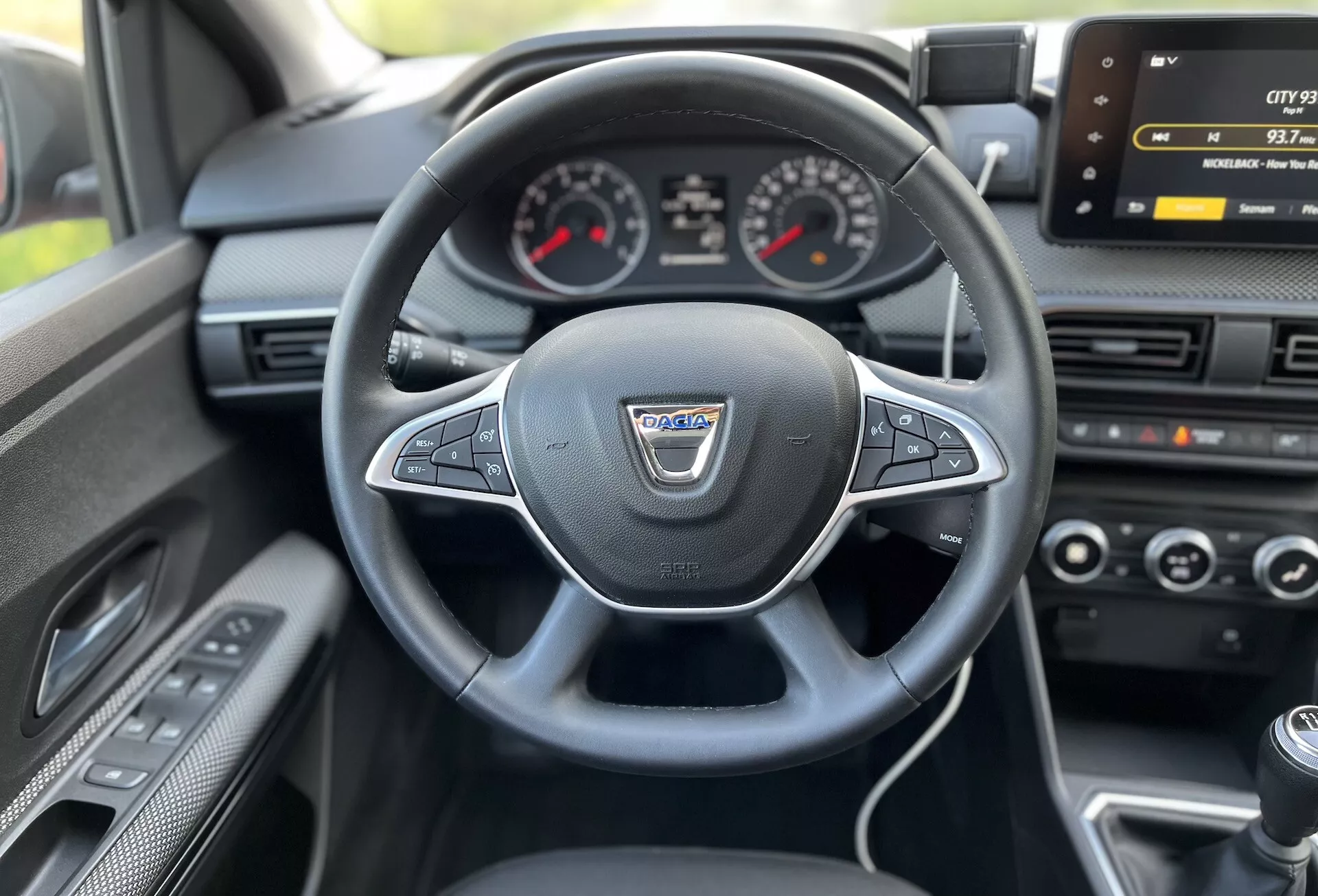 Vista interior de um Dacia Jogger 2022 do assento do motorista mostrando o volante multifuncional revestido de couro, conjunto de instrumentos analógico com display digital central, tela de infotenimento, controle de clima e alavanca de câmbio manual