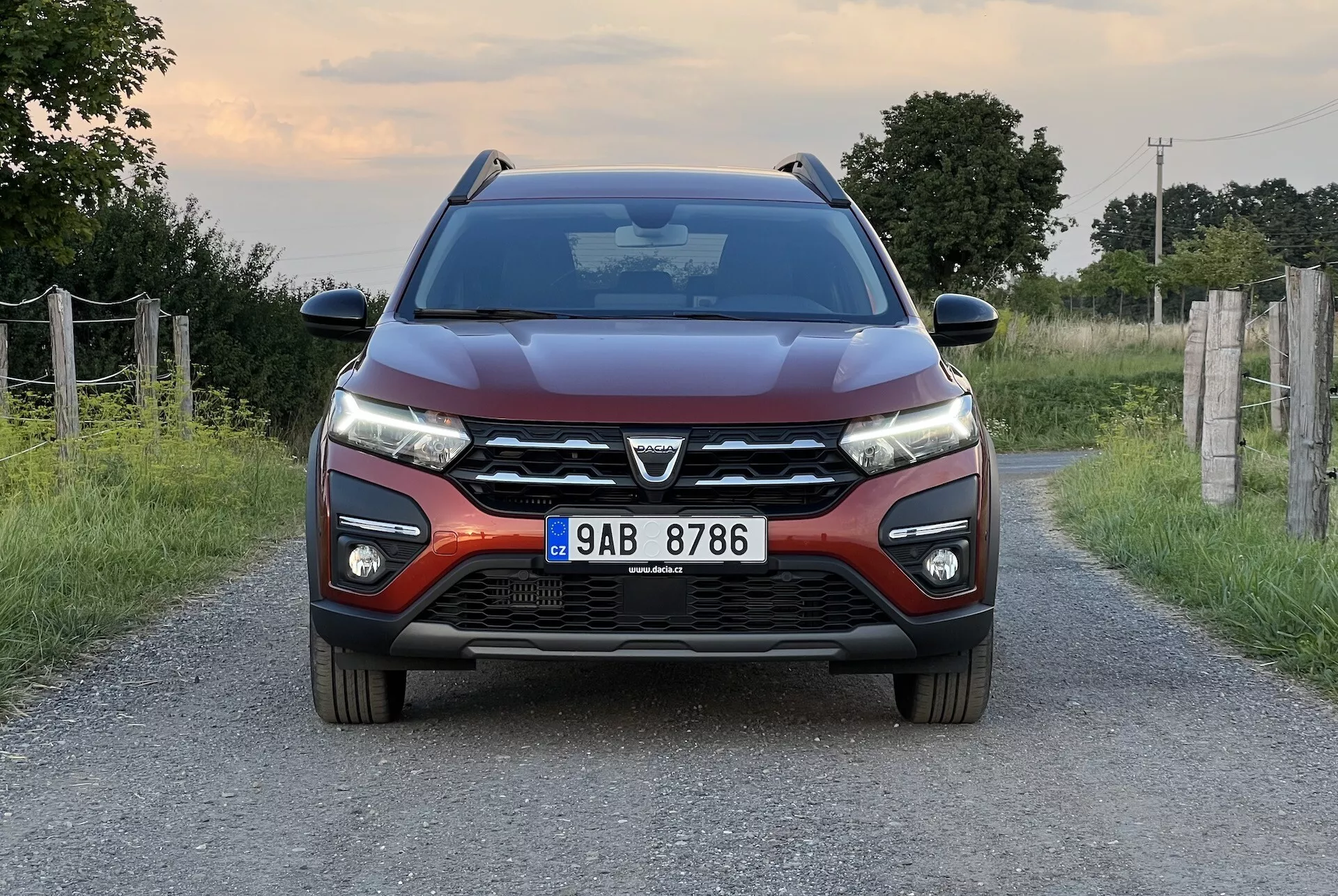 Dacia Jogger 2022 posicionado numa estrada rural estreita, apresentando uma vista frontal simétrica que destaca o design moderno do carro, incluindo uma grade frontal proeminente com um grande emblema, faróis LED elegantes, linhas contornadas no capô, para-choque dianteiro com faróis de neblina e sensores de estacionamento, e espelhos laterais com setas integradas.