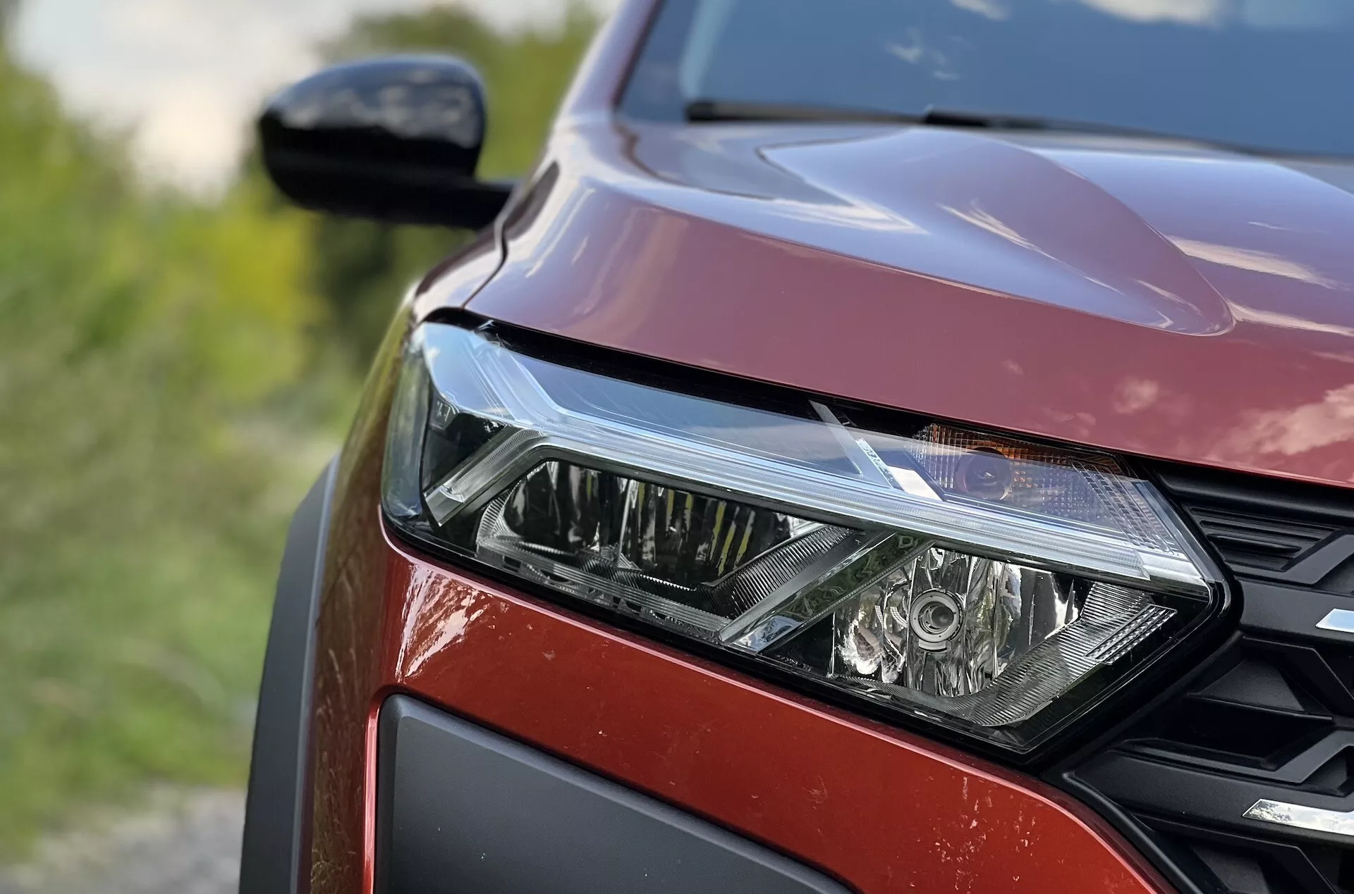 Vista de perto do lado frontal esquerdo do Dacia Jogger 2022 destacando o conjunto de faróis LED, padrão de grade hexagonal e acabamento de pintura vermelha metálica