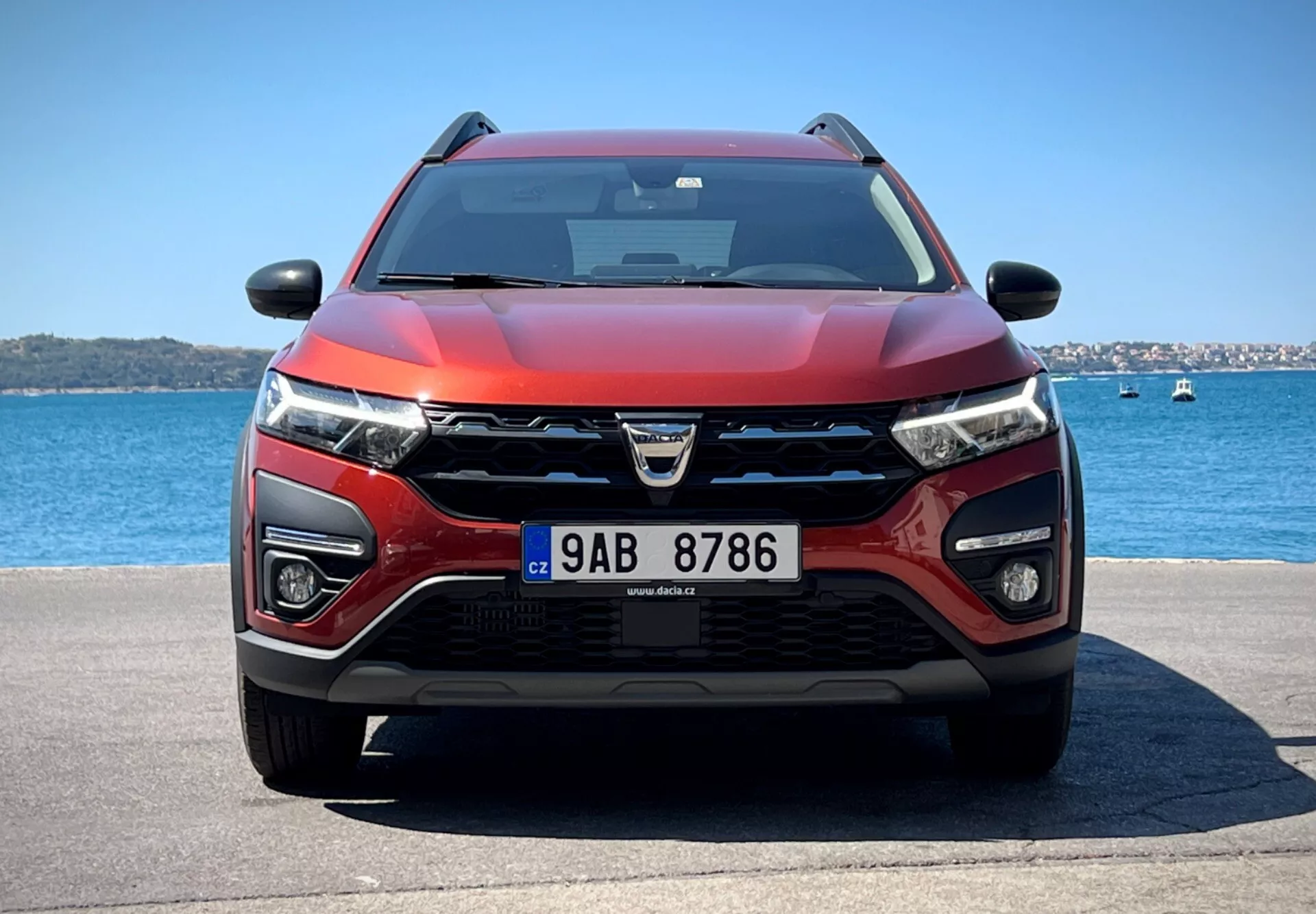 Dacia Jogger 2022 exibindo seu design moderno de crossover e características tecnológicas avançadas em um cenário aquático sereno, apresentando uma grade proeminente com logotipo, faróis de LED, faróis de neblina, potenciais sensores ADAS, entradas de ar para refrigeração e radar, esquema de cores vermelho e preto em duas tonalidades, capô aerodinâmico, equipado com limpadores de para-brisa indicando possível tecnologia sensível à chuva, retrovisores laterais com indicadores de direção integrados possivelmente contendo tecnologia de monitoramento de ponto cego, sob céus limpos.