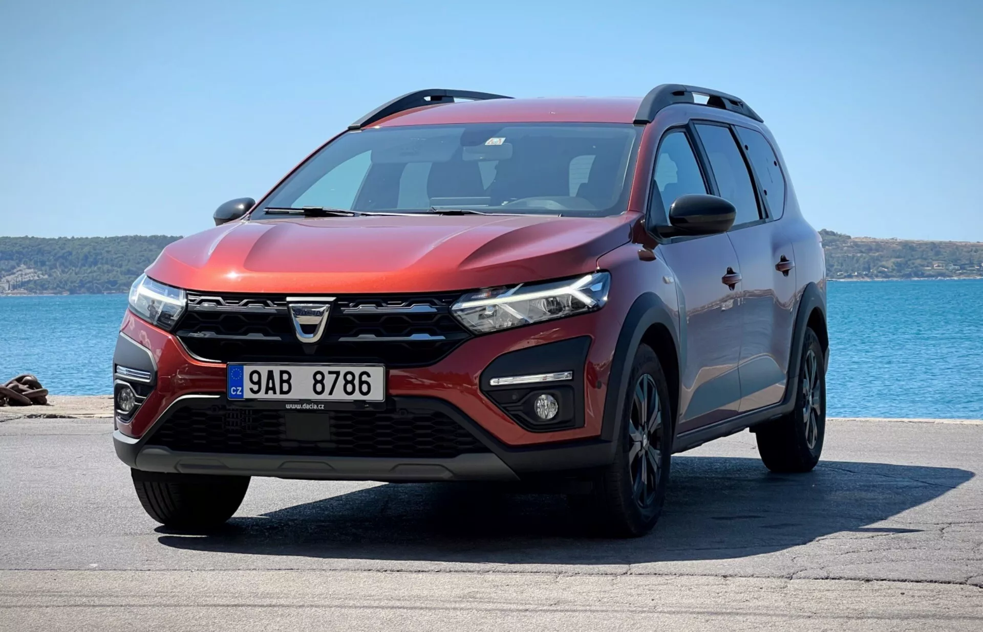 Visão frontal de três quartos do SUV Dacia Jogger 2022 estacionado com o fundo do oceano, mostrando design de grade distintivo, faróis LED, luzes de nevoeiro, para-choque dianteiro aerodinâmico com entradas de ar e sensores de estacionamento, contornos musculosos do capô, rodas de liga leve, sinais de volta integrados nos espelhos laterais, perfil lateral com proteção inferior, barras de teto, janelas com película, e exterior vermelho metálico sob luz solar brilhante indicando perfil de design, características tecnológicas e utilidade.