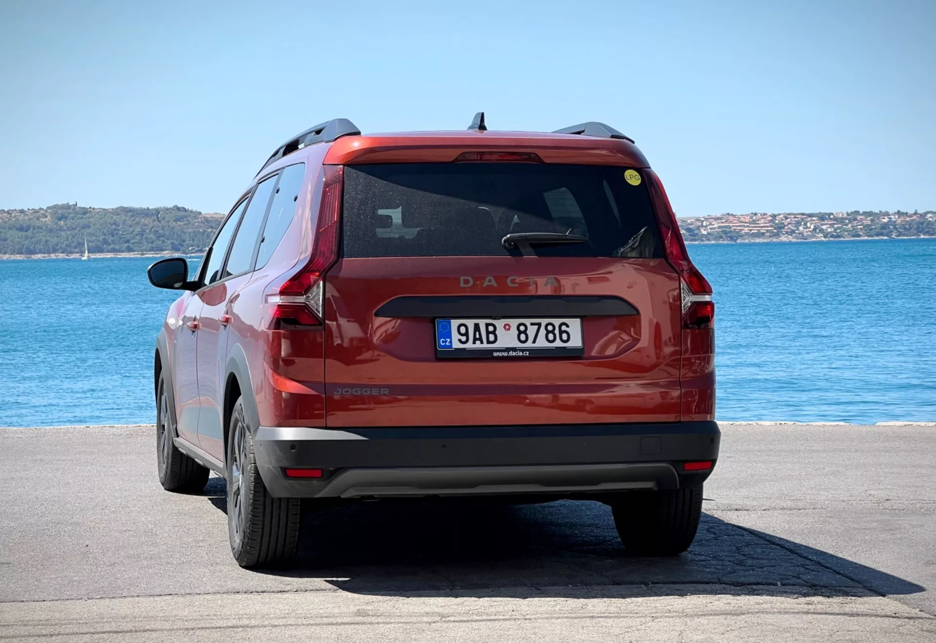Vista traseira de um SUV compacto vermelho Dacia Jogger 2022 estacionado numa área pavimentada junto à água sob a luz do dia, mostrando toda a parte traseira do veículo, incluindo as luzes traseiras, o vidro traseiro e a placa de matrícula contra um cenário de água azul e céus limpos.