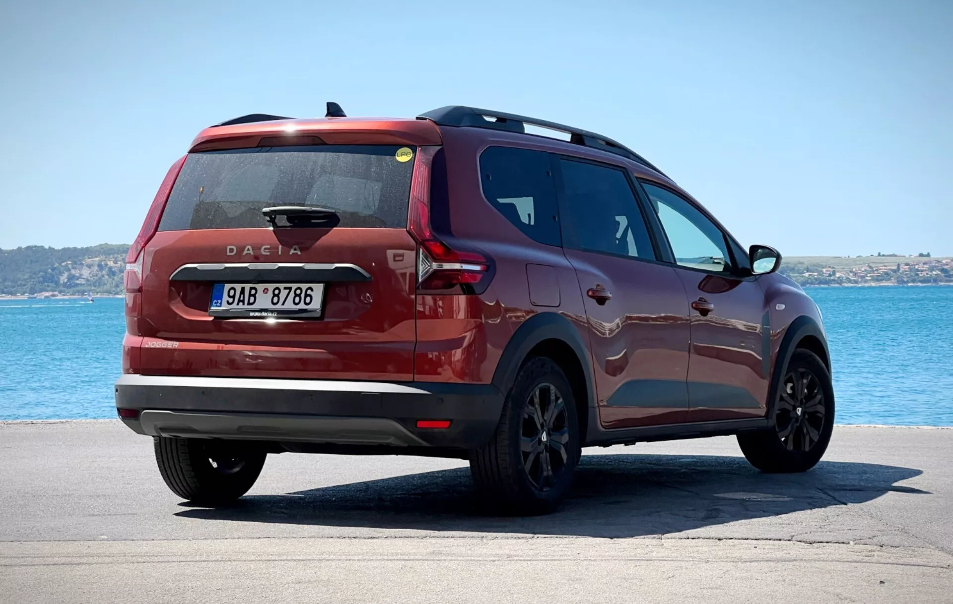 Dacia Jogger 2022 fotografada em uma vista traseira de três quartos posicionada em uma superfície pavimentada com um corpo de água e céu limpo ao fundo, exibindo luzes traseiras modernas, um distintivo da Dacia centralmente colocado, e um perfil lateral elegante contra um ambiente costeiro ou à beira do lago tranquilo.