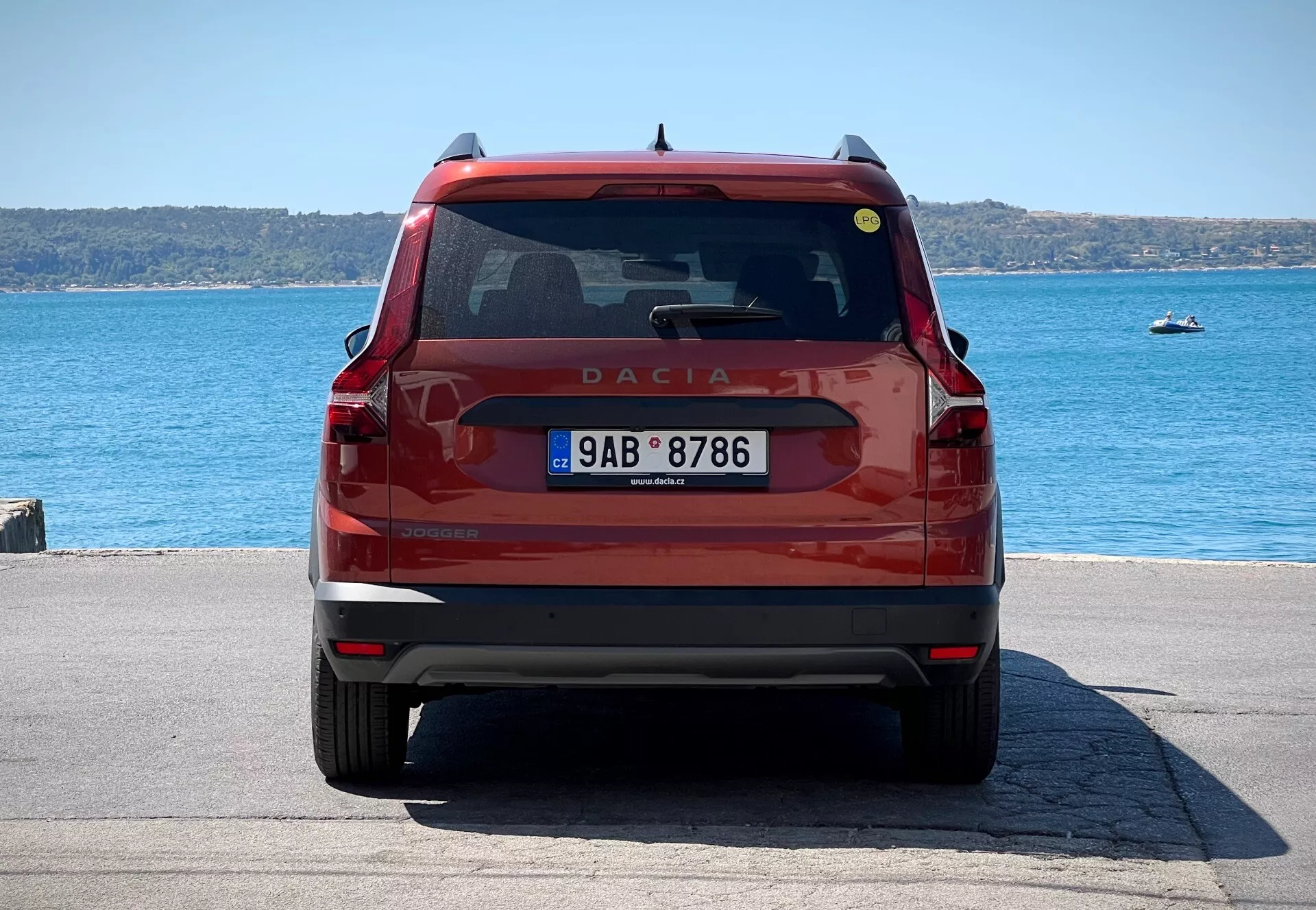 Vista traseira de um Dacia Jogger 2022 vermelho, mostrando porta-malas com emblema e nome do modelo, vidro traseiro com limpador e luz de freio, lanternas traseiras modernas, placa de licença e para-choque com refletores e possíveis sensores de estacionamento, estacionado à beira-mar à luz do dia.