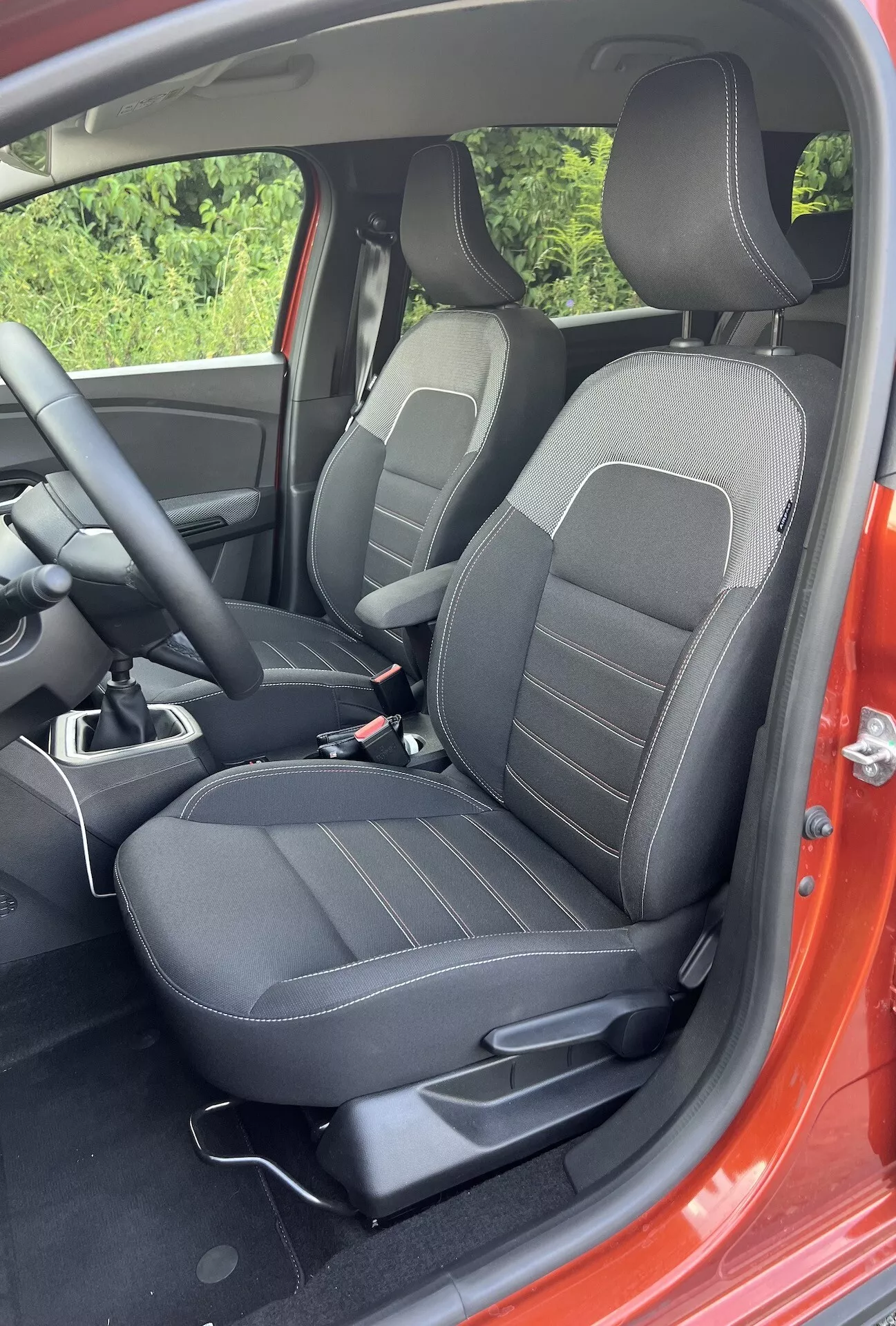 Vista detalhada do interior do Dacia Jogger 2022 mostrando a área do passageiro da frente com assentos estofados em preto com padrões de linhas, controles de ajuste, parte do console central, e um vislumbre do exterior vermelho