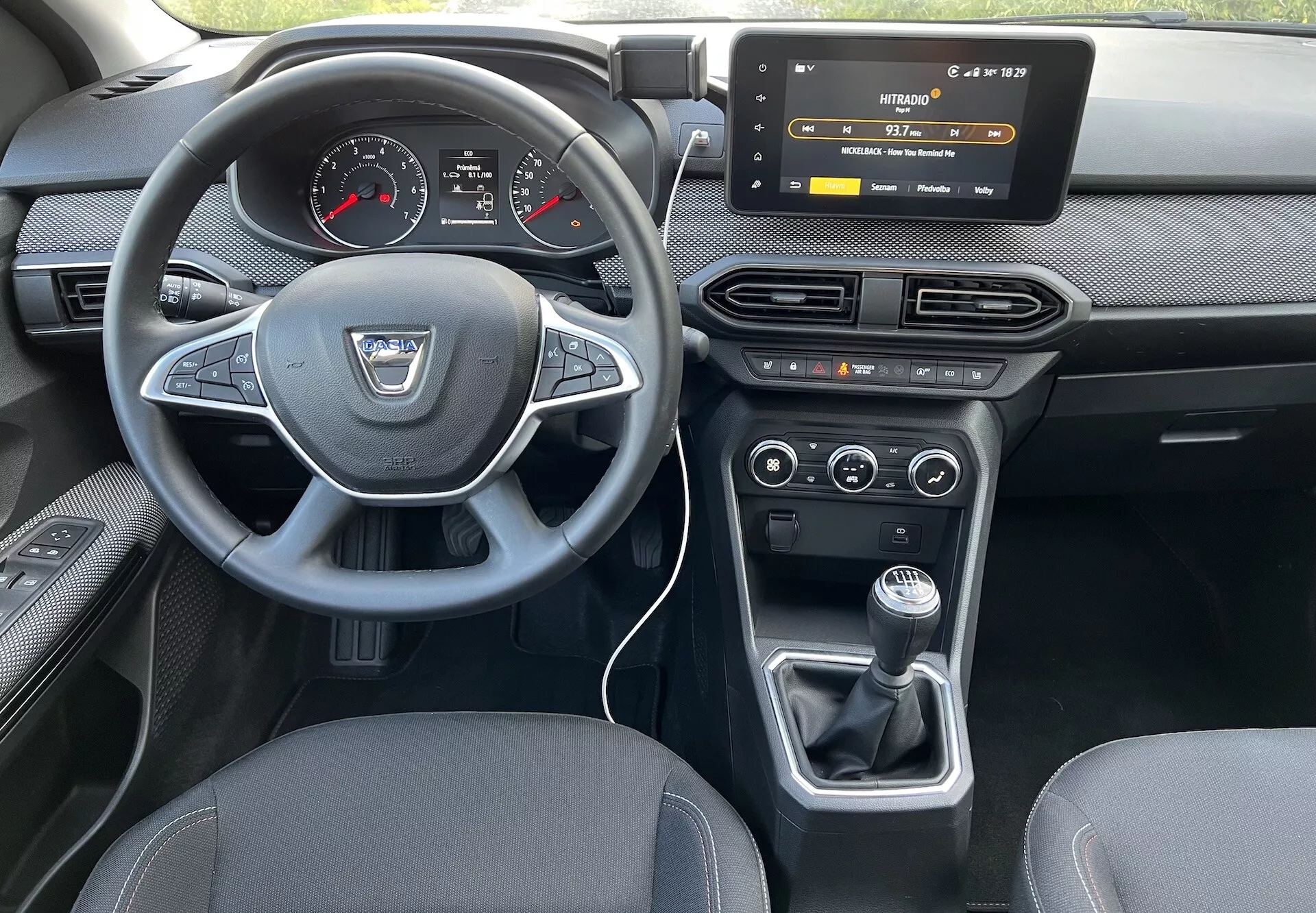 Vista interior de um Dacia Jogger 2022 a partir do assento traseiro, destacando o painel do condutor, volante com logo do fabricante e botões de controlo, cluster de instrumentos digital-analógico, sistema de infotenimento touchscreen, controles HVAC, controle de clima de duas zonas e alavanca de câmbio manual