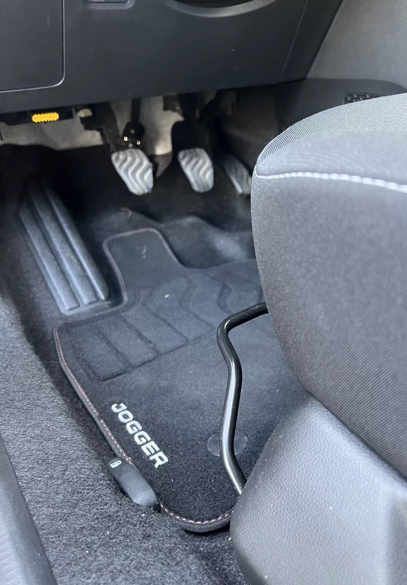 Interior do Dacia Jogger 2022 mostrando o espaço para os pés do lado do motorista com pedais metálicos, tapete de marca, assento de tecido cinza e acabamento em plástico preto