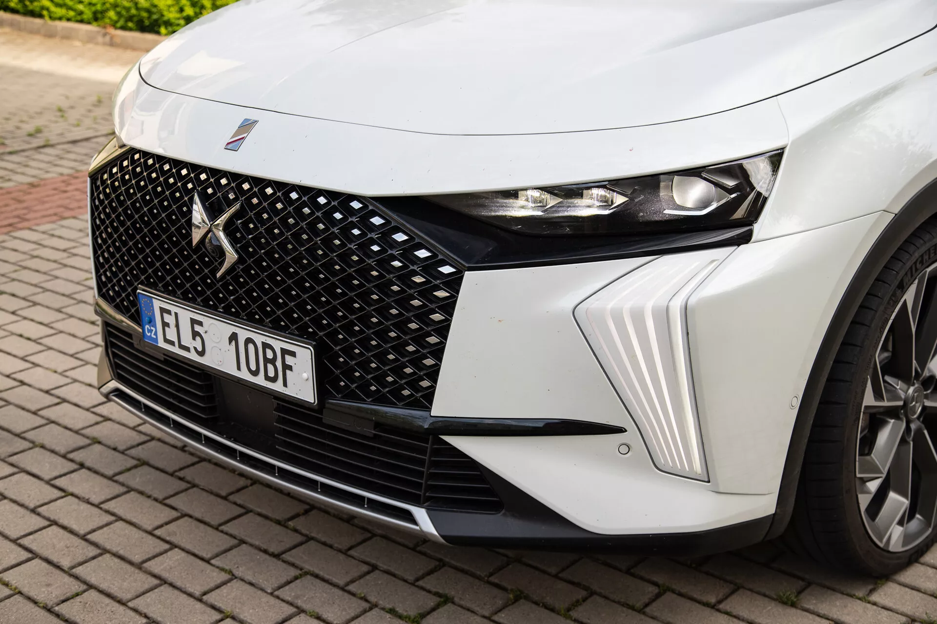 Visão detalhada do quadrante frontal esquerdo do DS Automobiles DS-7 2022 com foco detalhado no farol LED, design da grelha hexagonal com emblema central da marca, e contornos dinâmicos do capô junto com um vislumbre da roda de liga leve