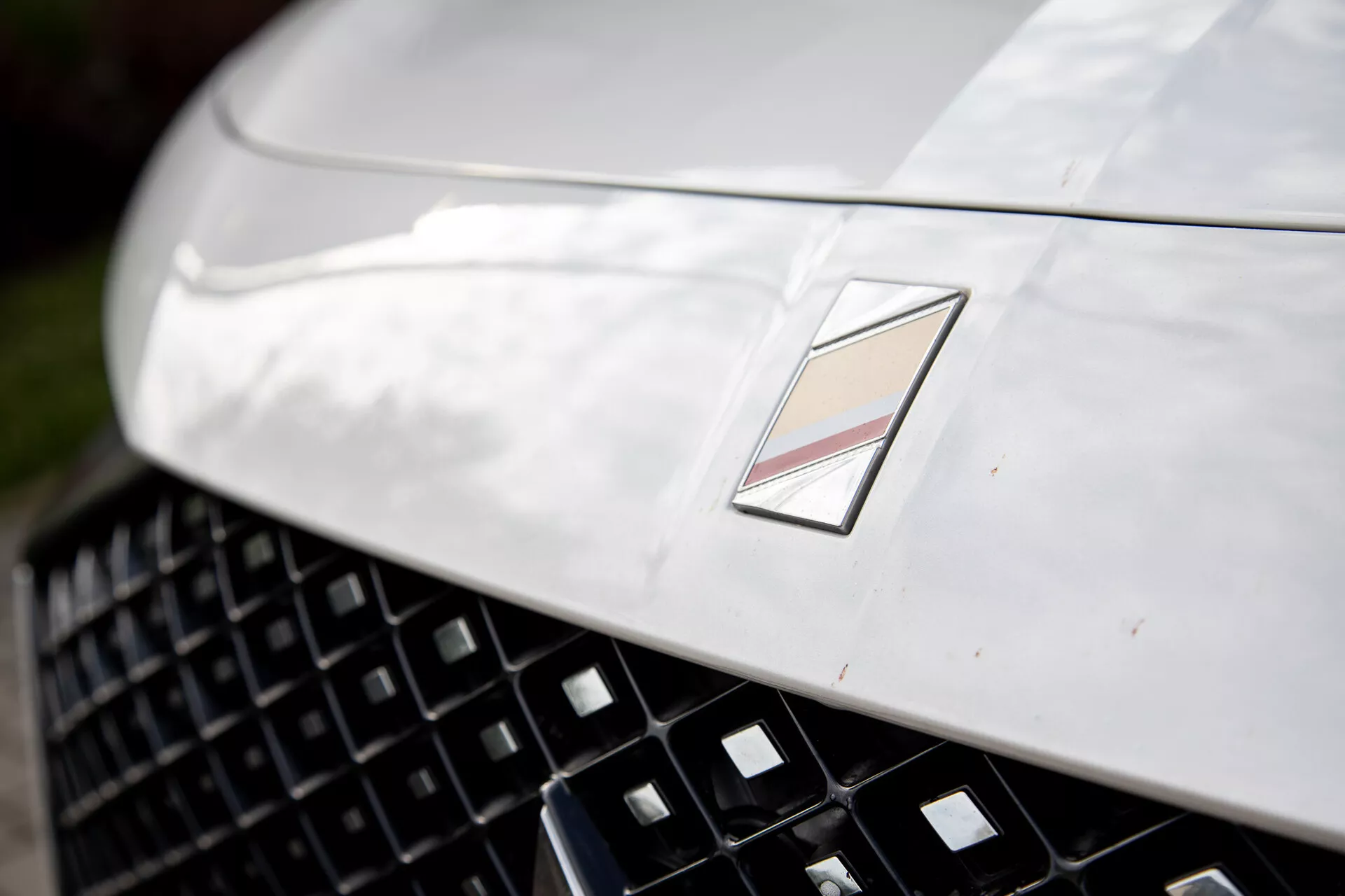 Vista de perto da seção frontal de um carro de luxo mostrando um capô metálico com design aerodinâmico, uma grade detalhada para melhor entrada de ar, e um emblema multifacetado significando o fabricante, sob reflexo da luz natural e profundidade de campo rasa
