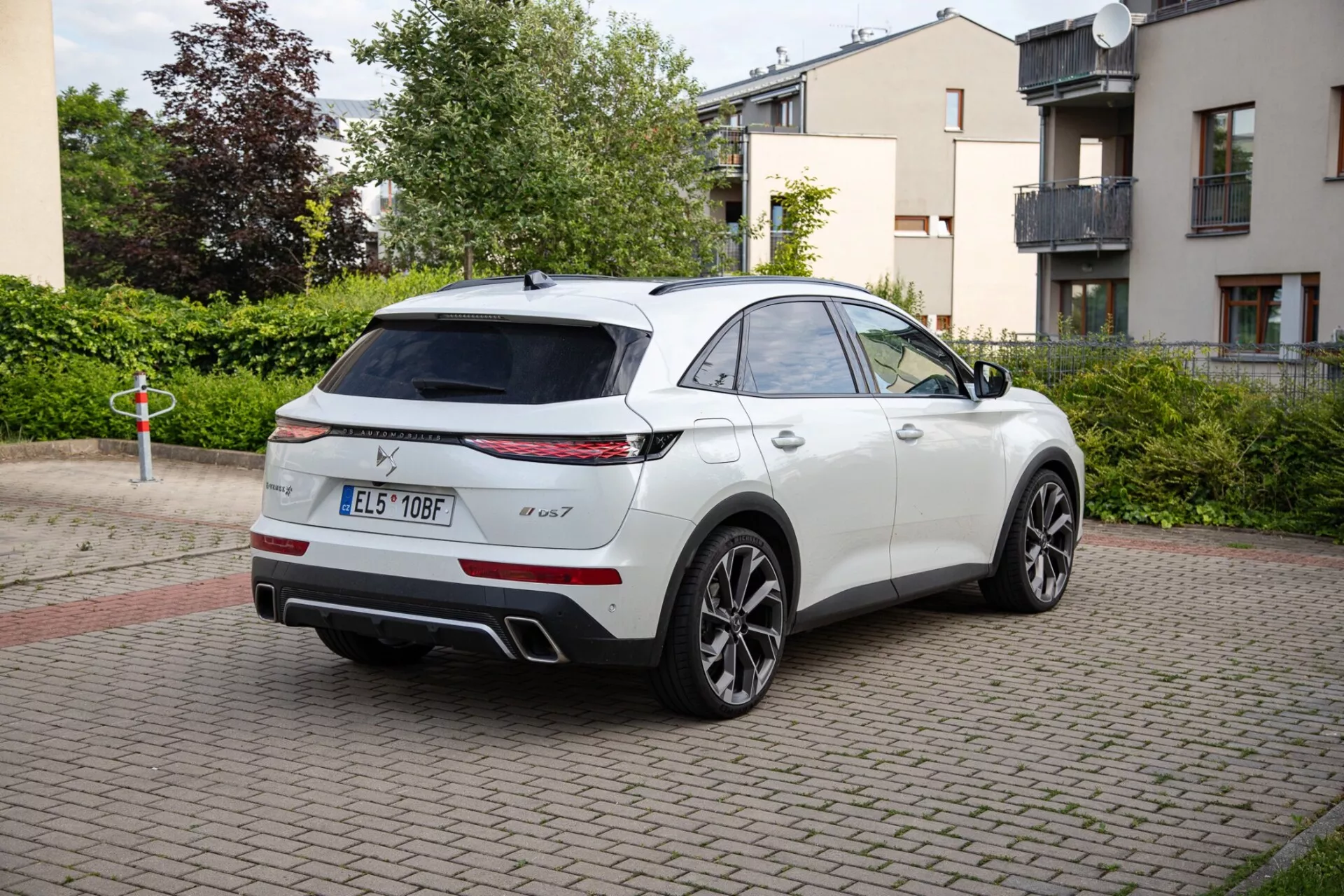 SUV branco DS Automobiles DS-7 de 2022 estacionado em uma área pavimentada com tijolos, mostrando lanternas traseiras LED, um sistema de escape duplo e rodas de liga leve esportivas. A imagem captura o design elegante do veículo, semelhante a um SUV coupé, de uma perspectiva traseira de três quartos, destacando um possível teto solar panorâmico e espelhos laterais com tecnologia avançada.