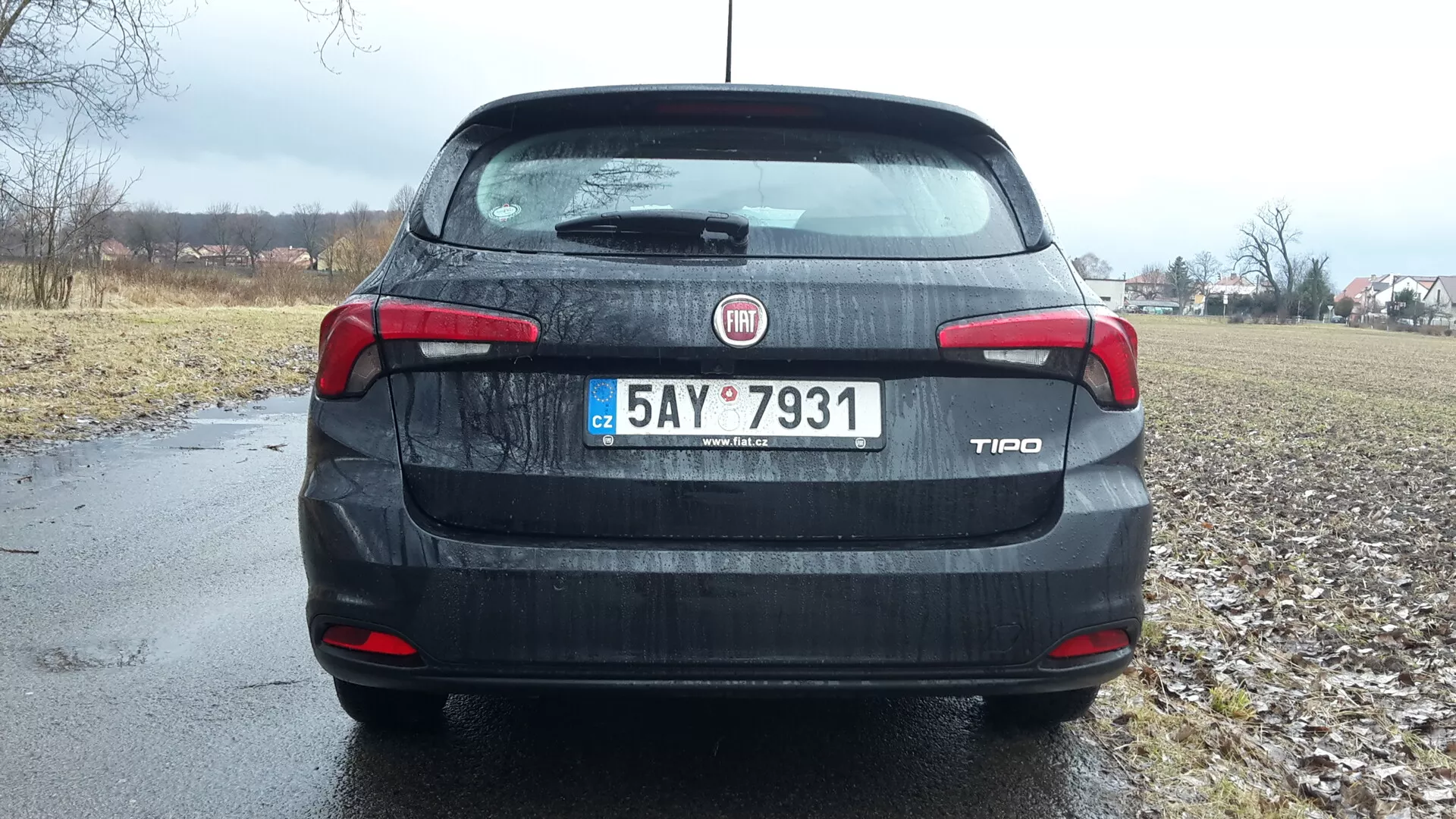 Vista traseira de um sedan Fiat Tipo 2016 sob céus nublados, apresentando identificação de marca e modelo no porta-malas, luzes traseiras e terceira luz de freio detalhadas, placa de registro e uma sugestão de ambiente rural ao fundo