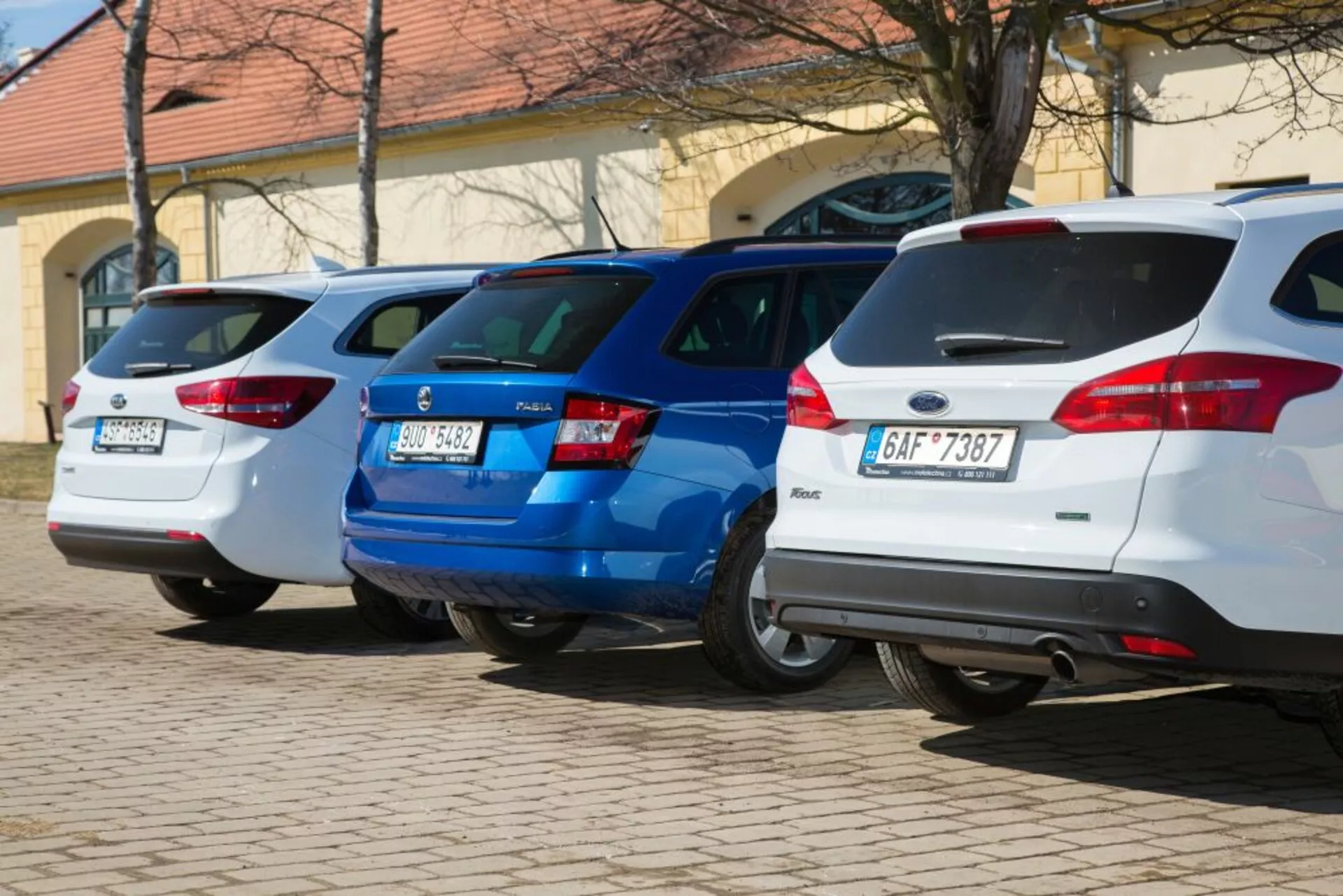 Uma linha de três hatchbacks, incluindo um Ford Focus branco de 2011, estacionados perpendicularmente com as extremidades traseiras voltadas para a câmera, exibindo luzes traseiras e características de design traseiro em uma área de estacionamento bem iluminada.