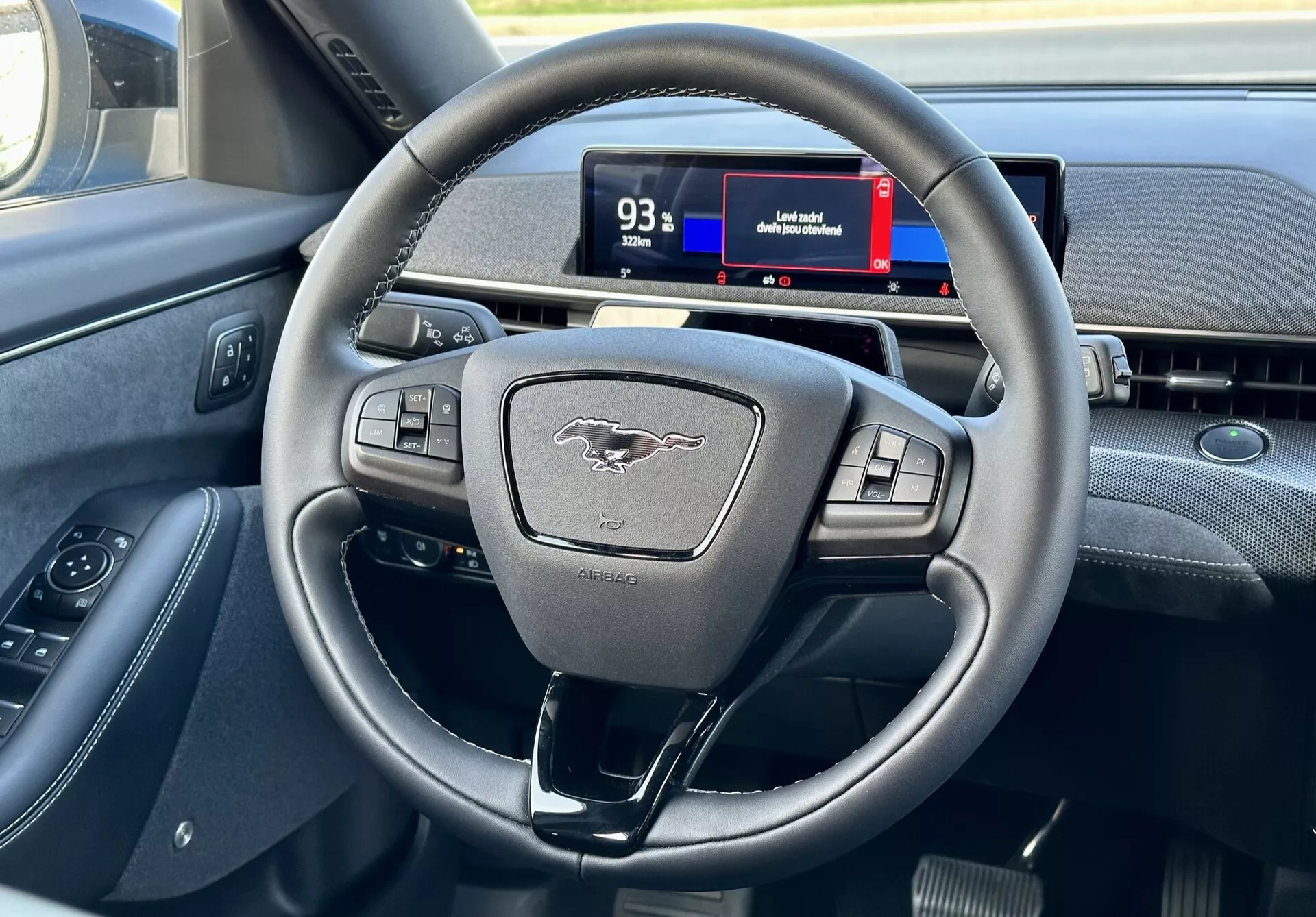 Interior do Ford Mustang Mach-E 2020 destacando o volante moderno de três raios com botões multifuncionais e o cluster de instrumentos digital da perspectiva do motorista, mostrando controles, emblema e dicas de navegação.