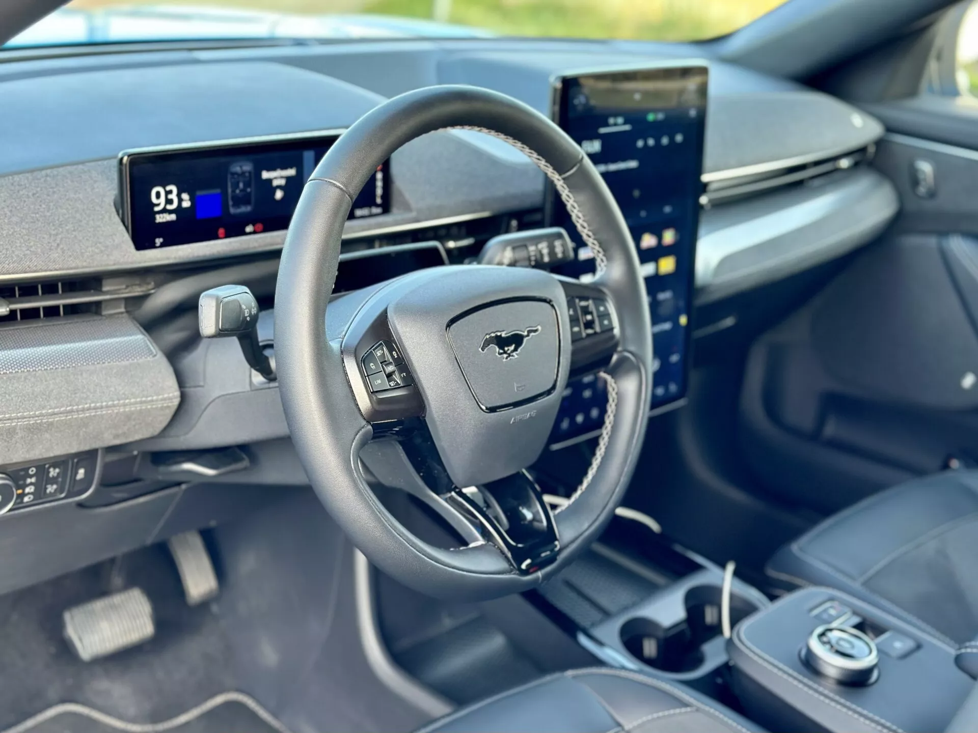 Vista interior de um Ford Mustang Mach-E 2020 focada no cockpit do motorista pela perspectiva do passageiro do banco da frente, apresentando um volante com emblema Mustang, painel digital em modo Parque mostrando autonomia de 93 milhas, e uma tela sensível ao toque central verticalmente orientada integrada com capacidades avançadas de infotainment.