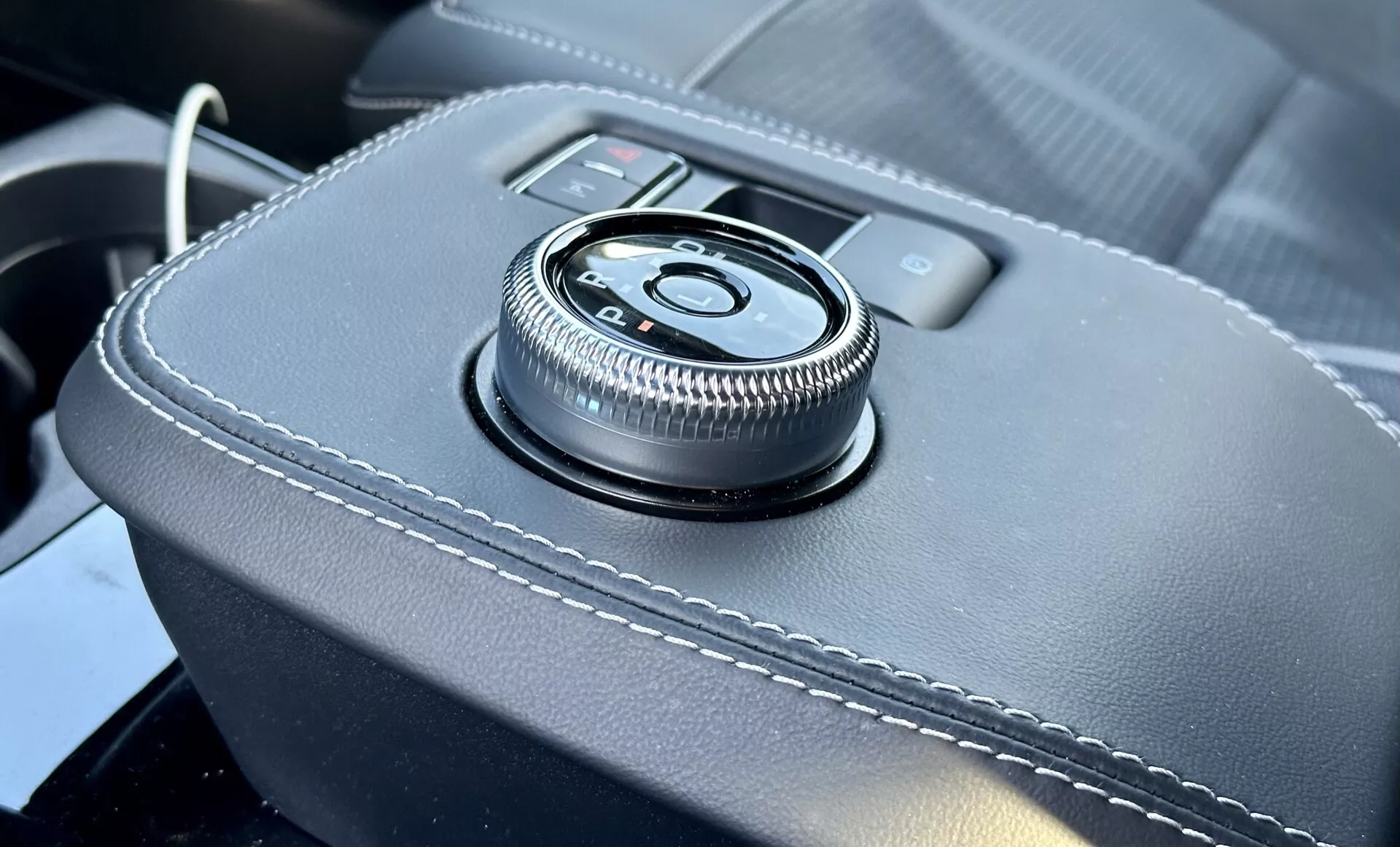 Vista detalhada do interior do Ford Mustang Mach-E 2020 com um controlo rotativo na consola central em uma superfície revestida a couro com costuras brancas, fornecendo uma interface ergonómica para o sistema de infotenimento