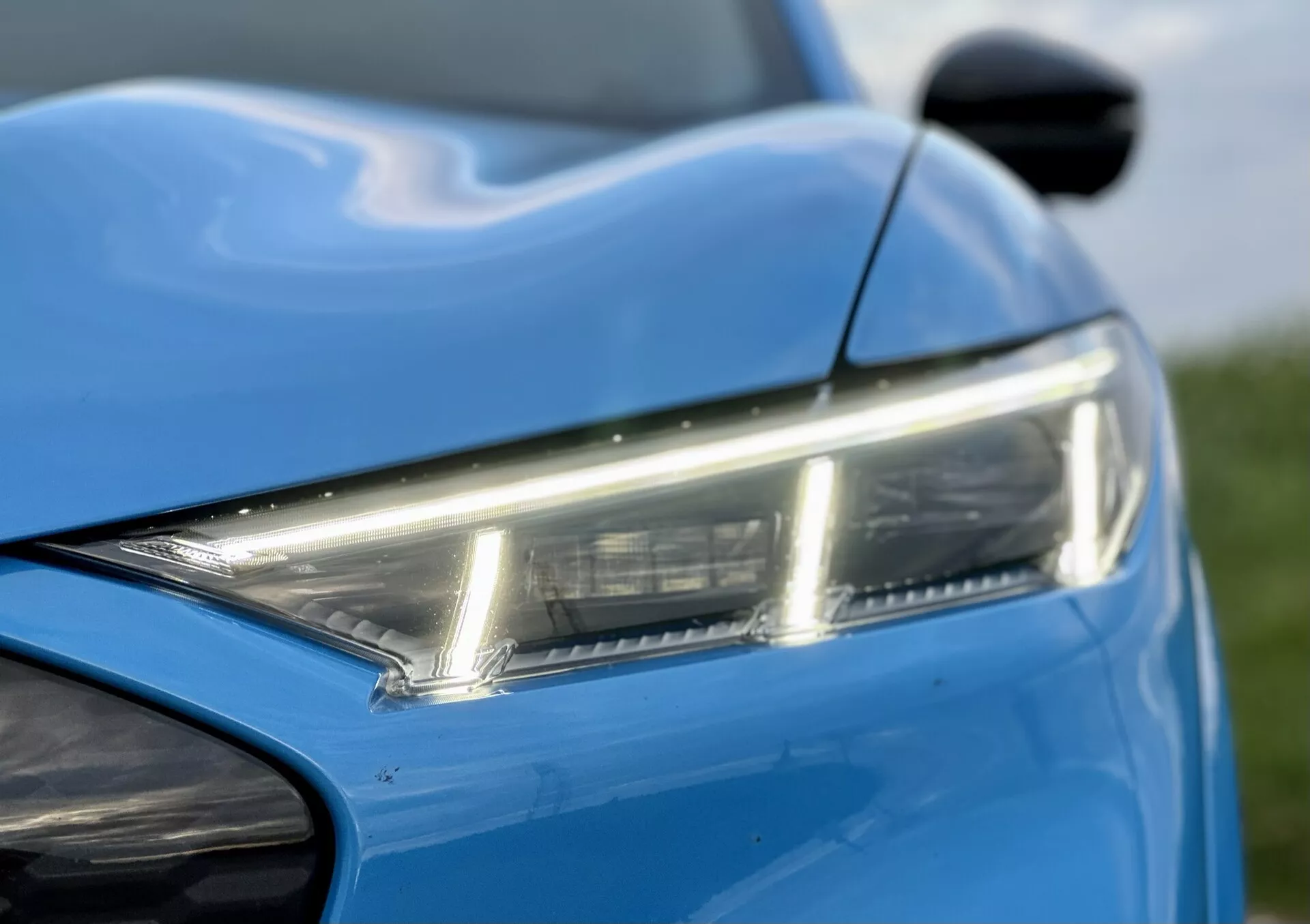Visão detalhada da seção frontal esquerda do Ford Mustang-Mach-E 2020, destacando o farol LED elegante, a pintura azul vibrante e as linhas dinâmicas da carroceria, com uma visão do espelho lateral e do para-choque dianteiro, exibindo o design moderno e a estética.