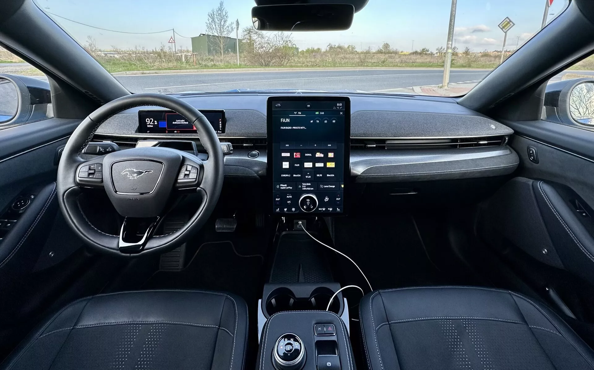 Vista da primeira pessoa do interior do Ford Mustang Mach-E 2020, mostrando o volante posicionado à esquerda com design de fundo plano e emblema, painel de instrumentos digital, grande sistema de infotenimento com tela vertical no console central com botões de controle do clima abaixo, portas USB, e estofamento de luxo em tons escuros com costura detalhada, indicando características avançadas de segurança e assistência, com um fundo exterior rural visível através do pára-brisa.