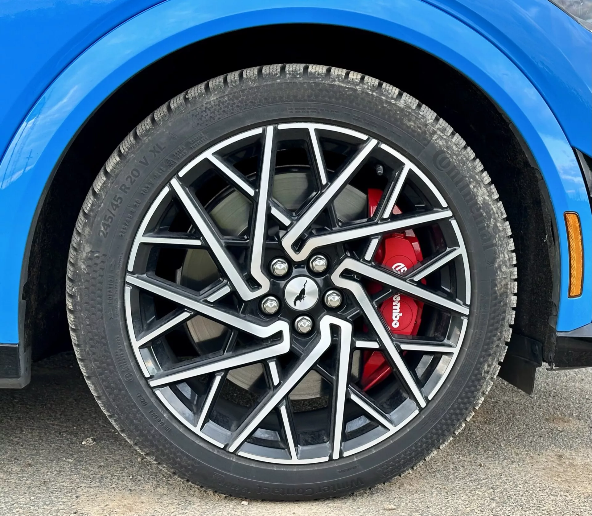 Close-up da roda dianteira direita do Ford Mustang Mach-E 2020 com aro de liga leve multiraiado e sistema de freio de alto desempenho com pinça vermelha, exibindo detalhes do pneu para todas as estações e um segmento do para-lama azul vivo, tirada de um ângulo baixo.