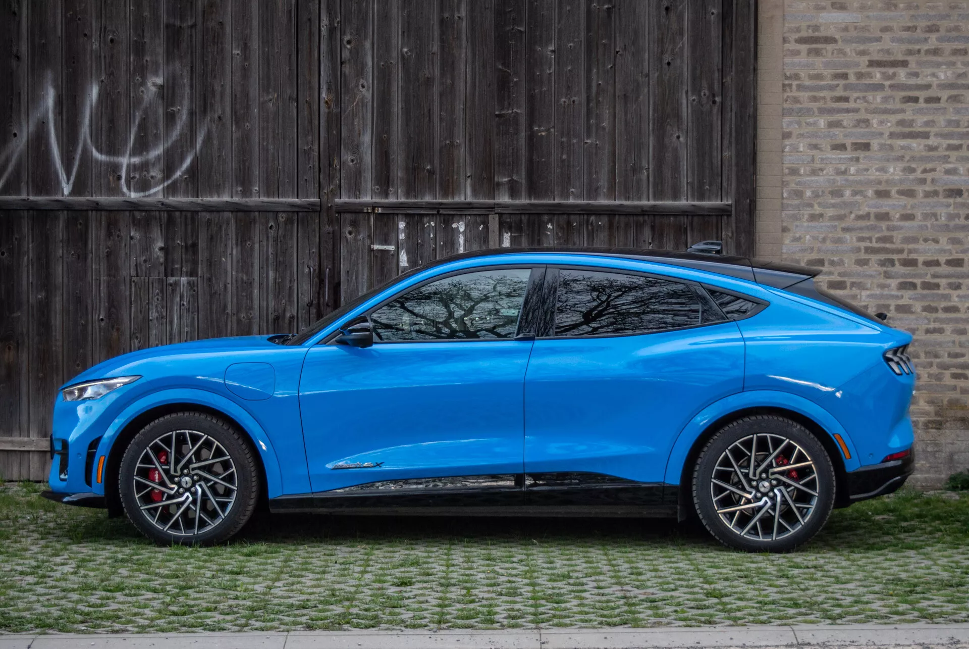SUV crossover azul Ford Mustang Mach-E 2020 estacionado paralelamente a uma porta de garagem de madeira rústica com grafite, visão lateral destacando design aerodinâmico, linha do teto inclinada estilo cupê, modelo de quatro portas com maçanetas integradas, espelhos laterais estilizados aerodinamicamente com sinais de direção, grandes rodas de liga leve em dois tons com pinças de freio vermelhas, altura moderada do solo, carroceria contornada com linhas esculpidas e arcos de roda pronunciados, linha de janelas afunilada com pequena janela de quarto após o pilar C, e saias laterais esportivas